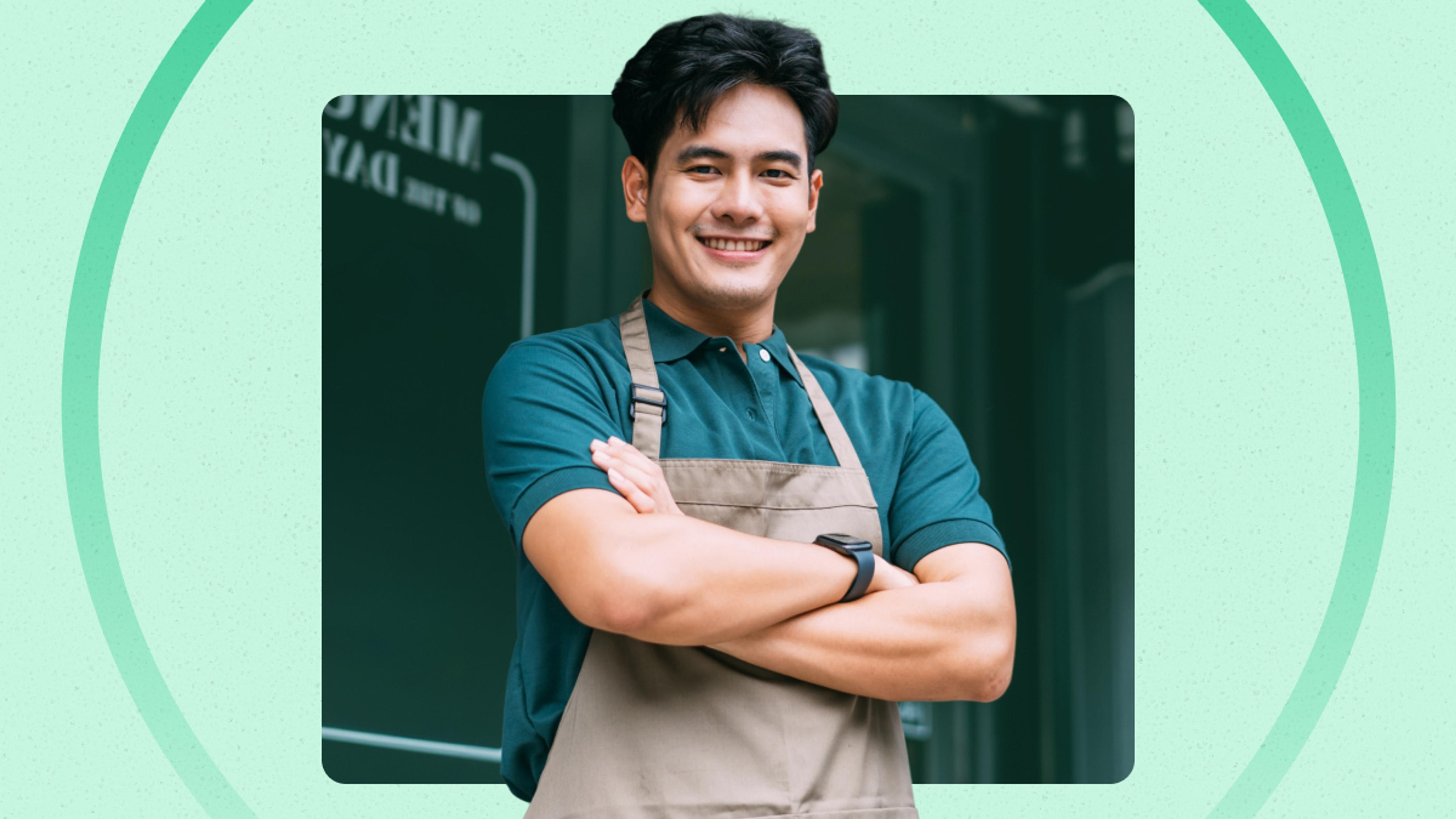 A man wearing a green polo shirt and beige apron stands with arms crossed, smiling in front of a dark green background.