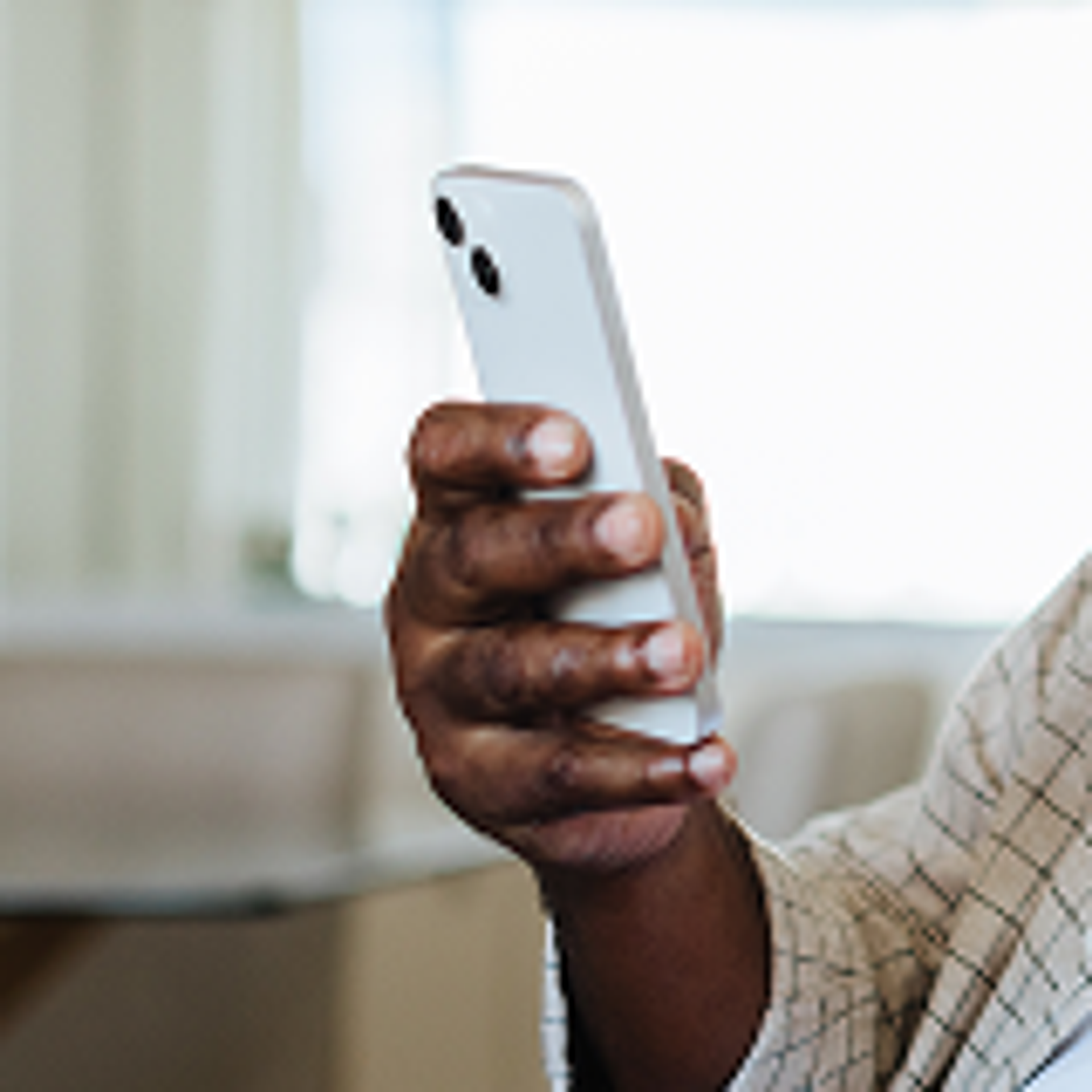 A person holding a white smartphone in one hand, with the screen facing away from the camera. The background is softly blurred, and the person is wearing a light-colored, plaid-patterned shirt.
