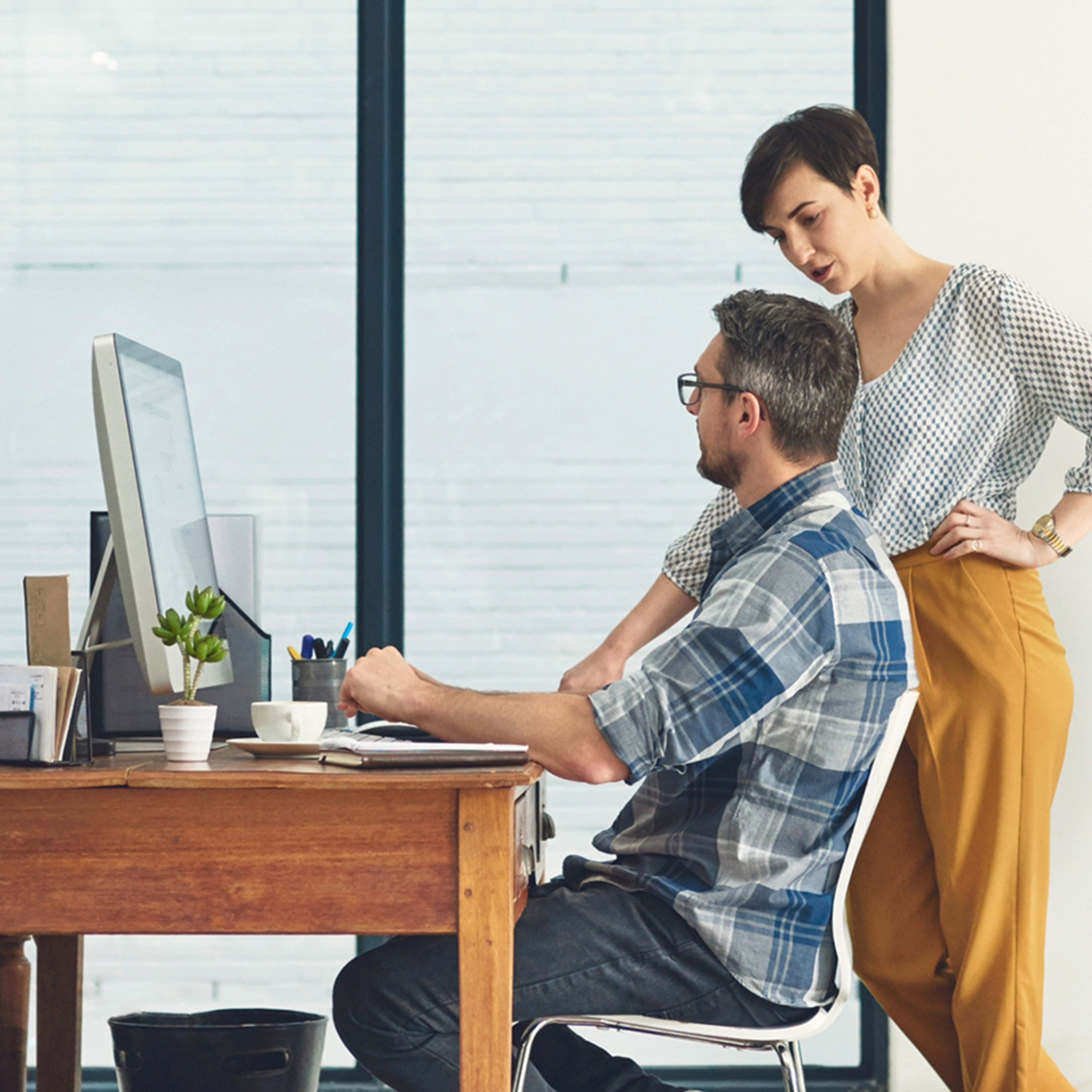 A man seated and working at a desk with a woman standing next to him.