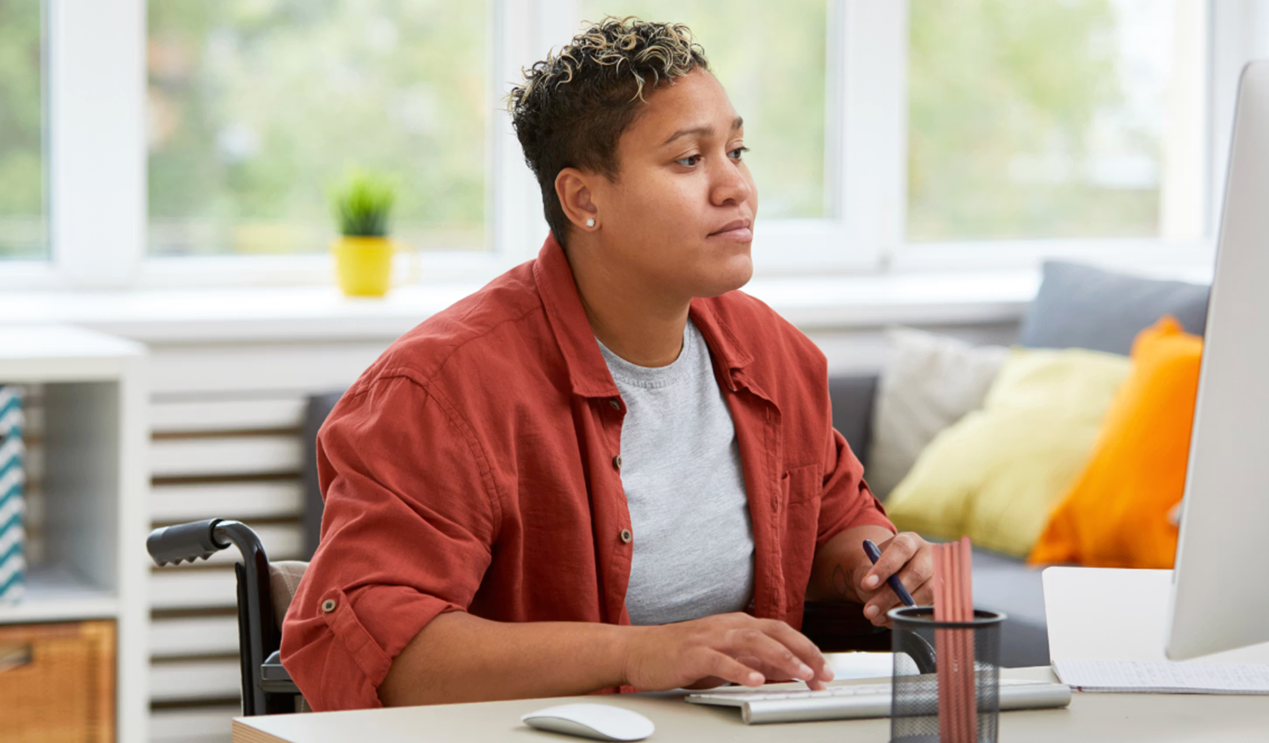 A person seated in a wheelchair works at a desk, looking at a computer monitor while typing and holding a pen. The setting appears to be a bright, modern home or office space with large windows and soft furnishings.