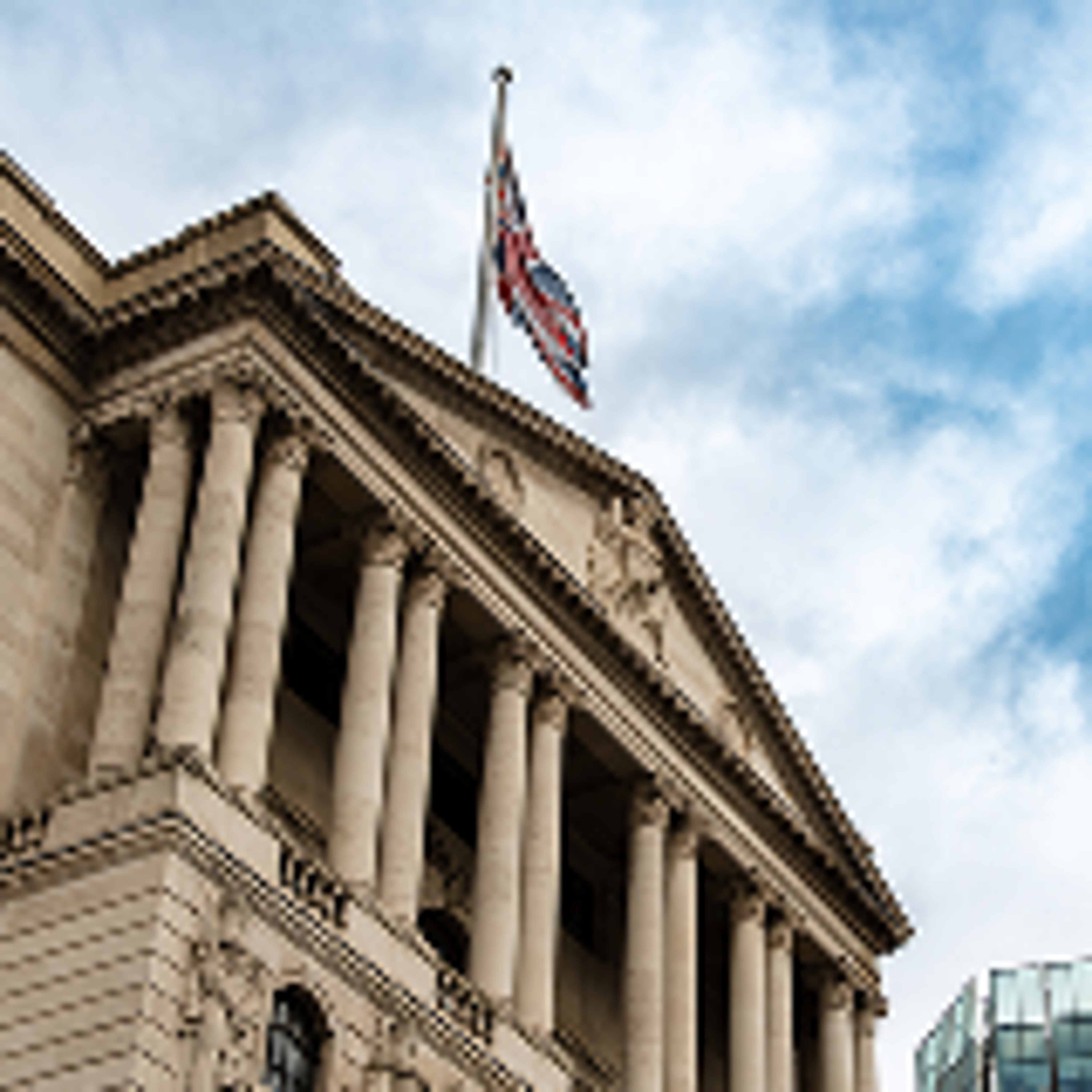An American flag flies at half-mast atop a classical building with columns, viewed from below against a partly cloudy sky.