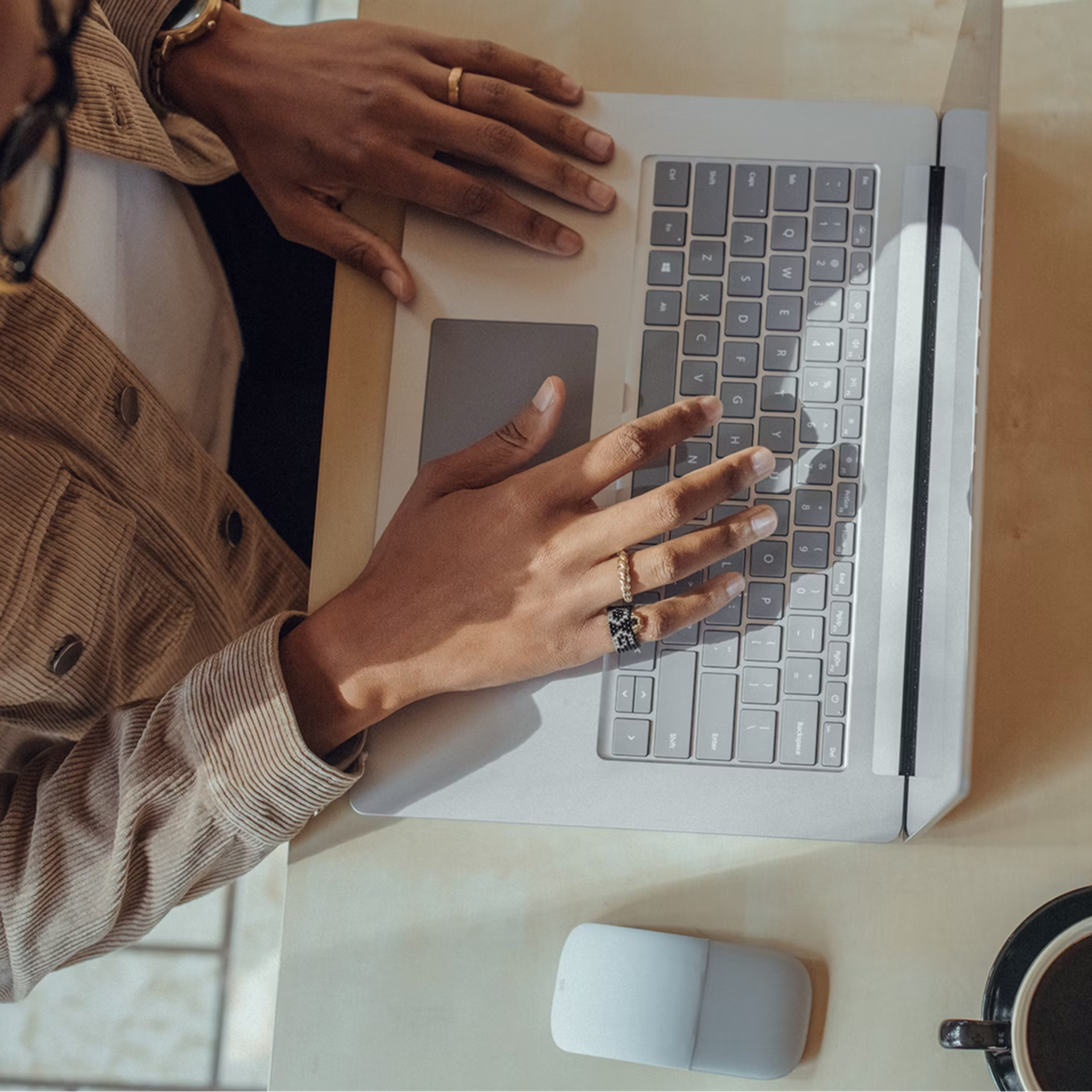 Hands using a laptop on a desk with a mouse and cup of coffee nearby.