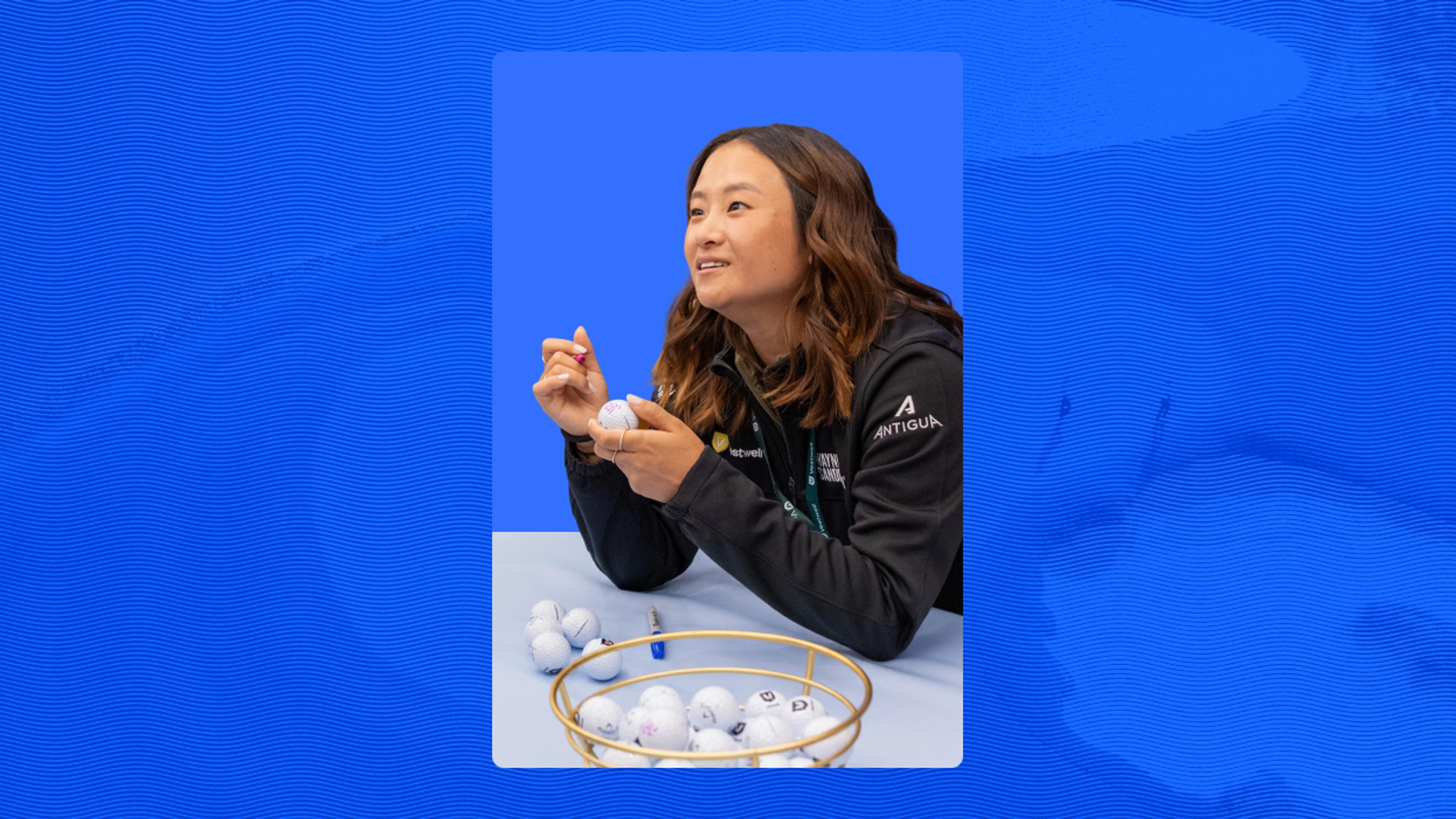 A person in a dark jacket sits at a table, holding a golf ball and marker, with a bowl of golf balls in front of them against a blue background.