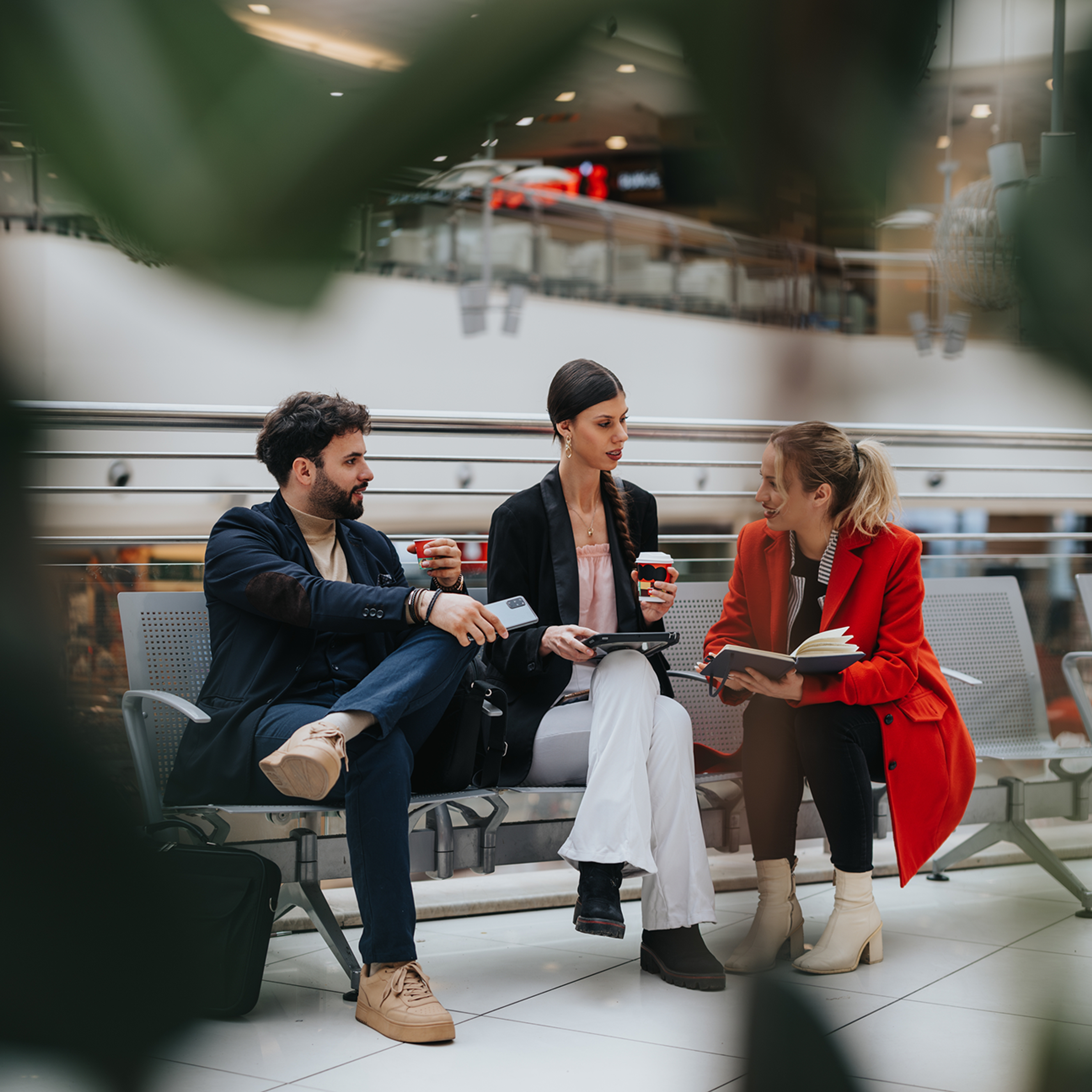 Three young adults sit on benches in a mall, engaged in conversation. One holds a smartphone, one has a coffee cup, and another takes notes in a notebook. Green leaves partially frame the scene.