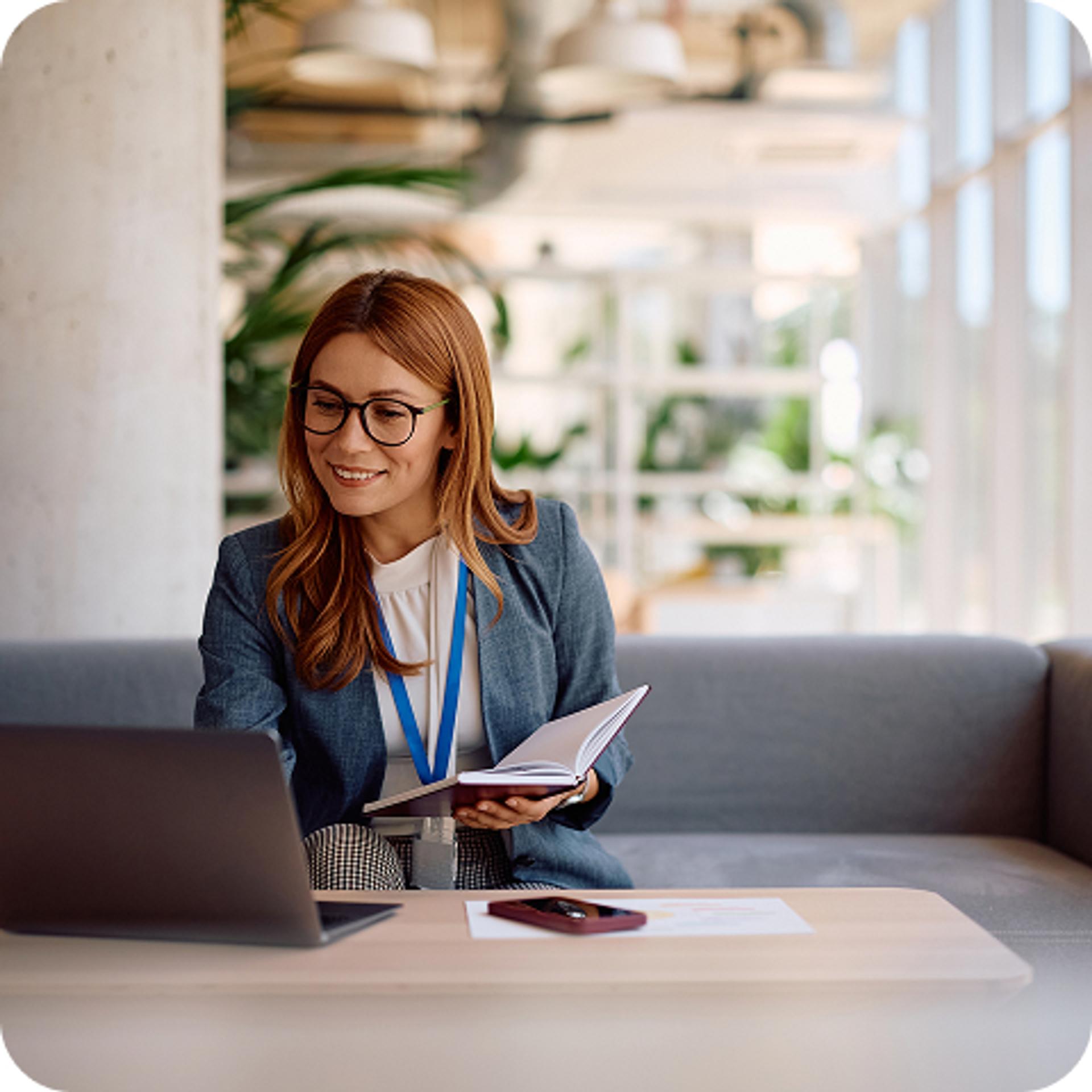 A woman wearing glasses and holding a notebook open as she works on a computer. 