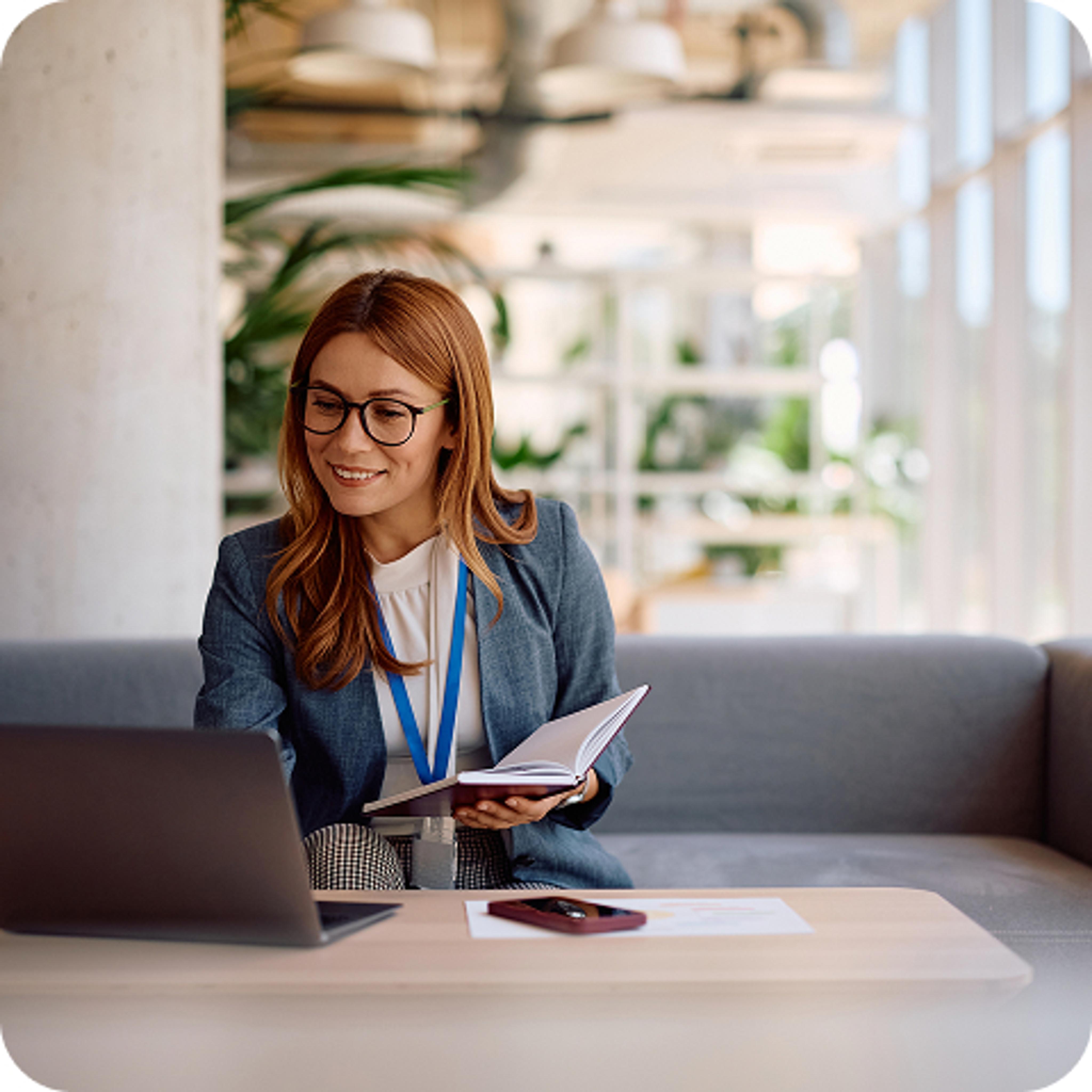 A woman wearing glasses and holding a notebook open as she works on a computer. 