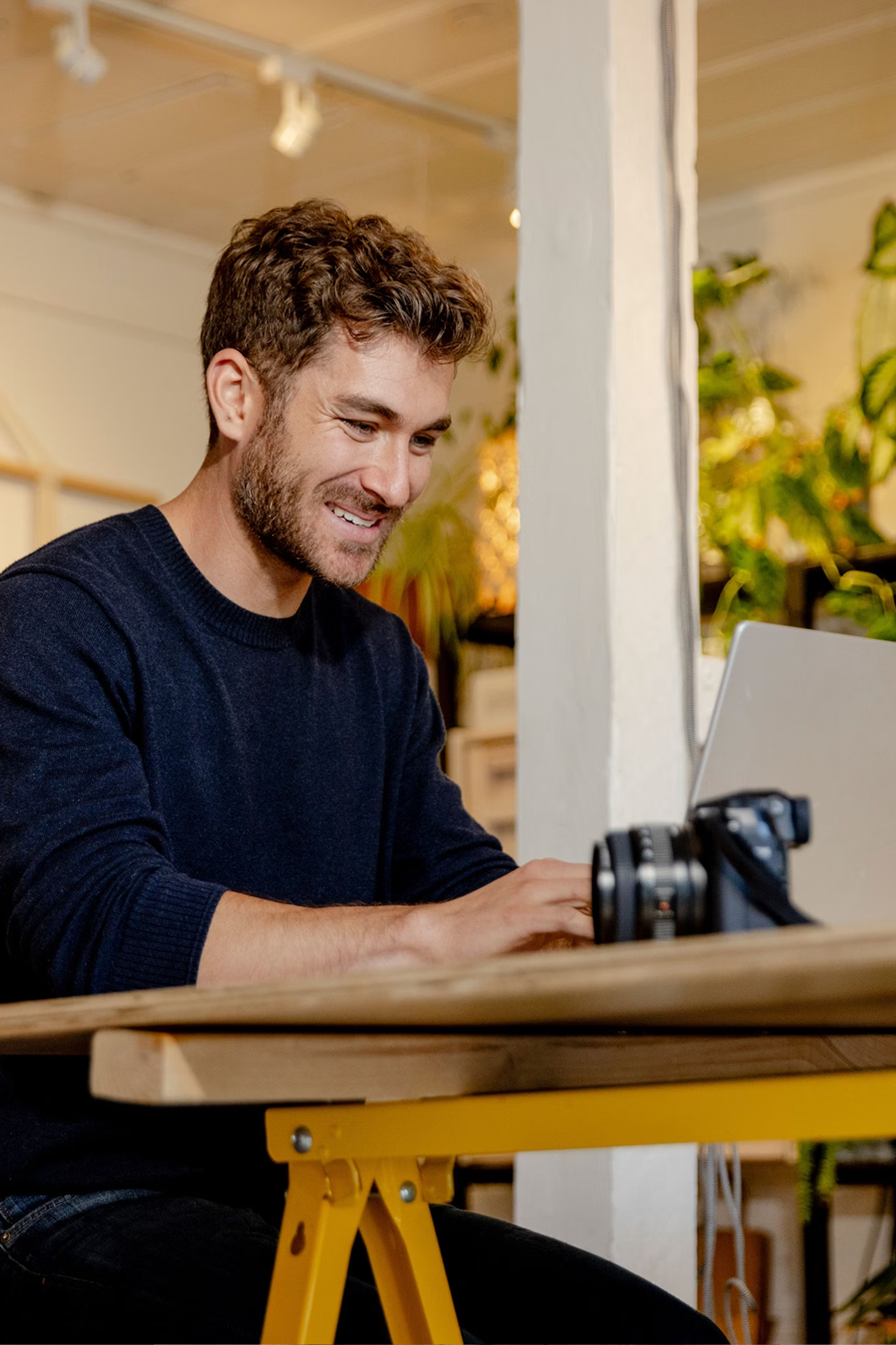 Person smiling while working on a laptop at a desk, with a camera nearby.