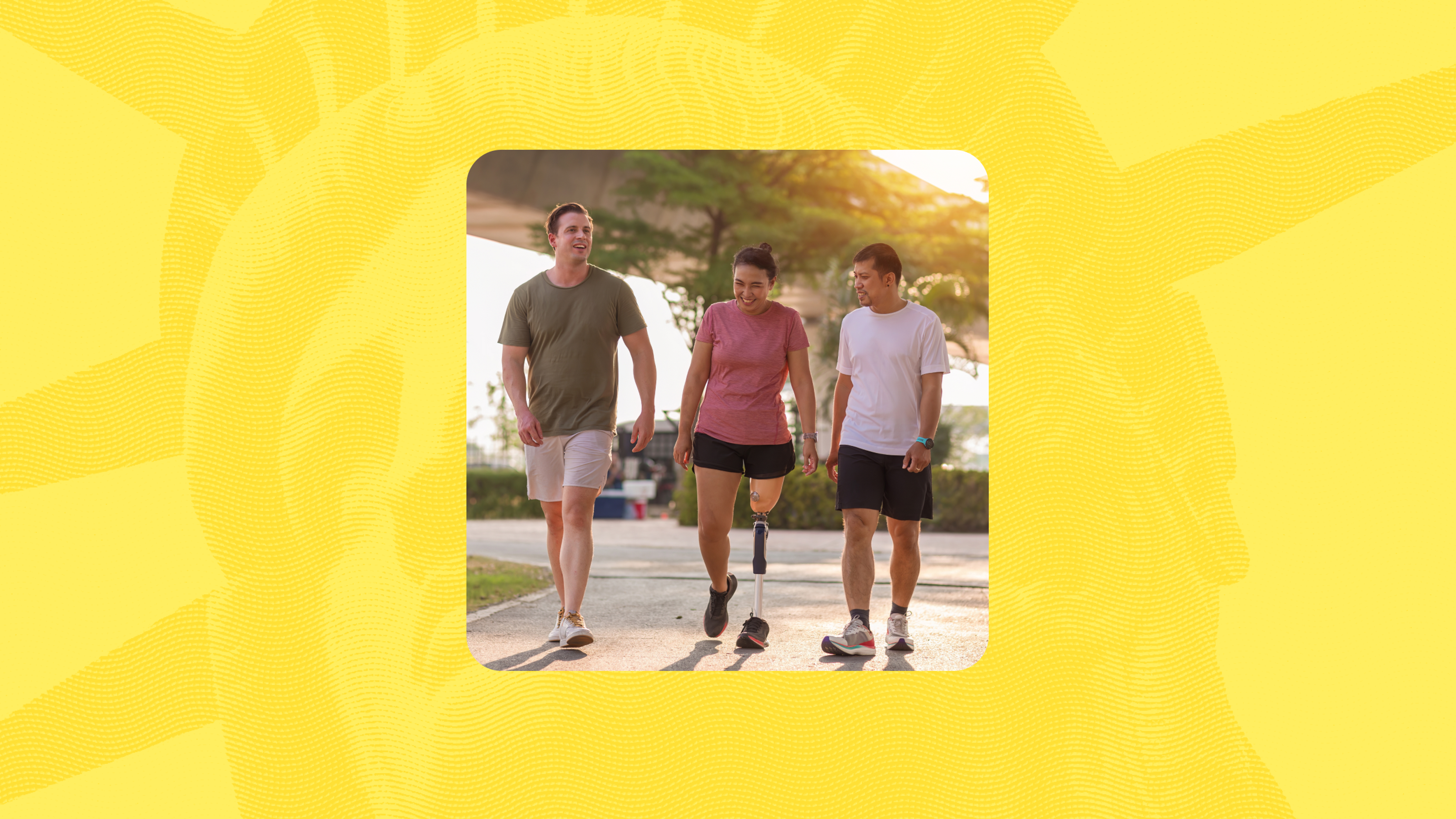 Three people walking together outdoors on a sunny day, smiling and talking; the person in the center uses a prosthetic leg, and all are dressed in casual athletic clothing.