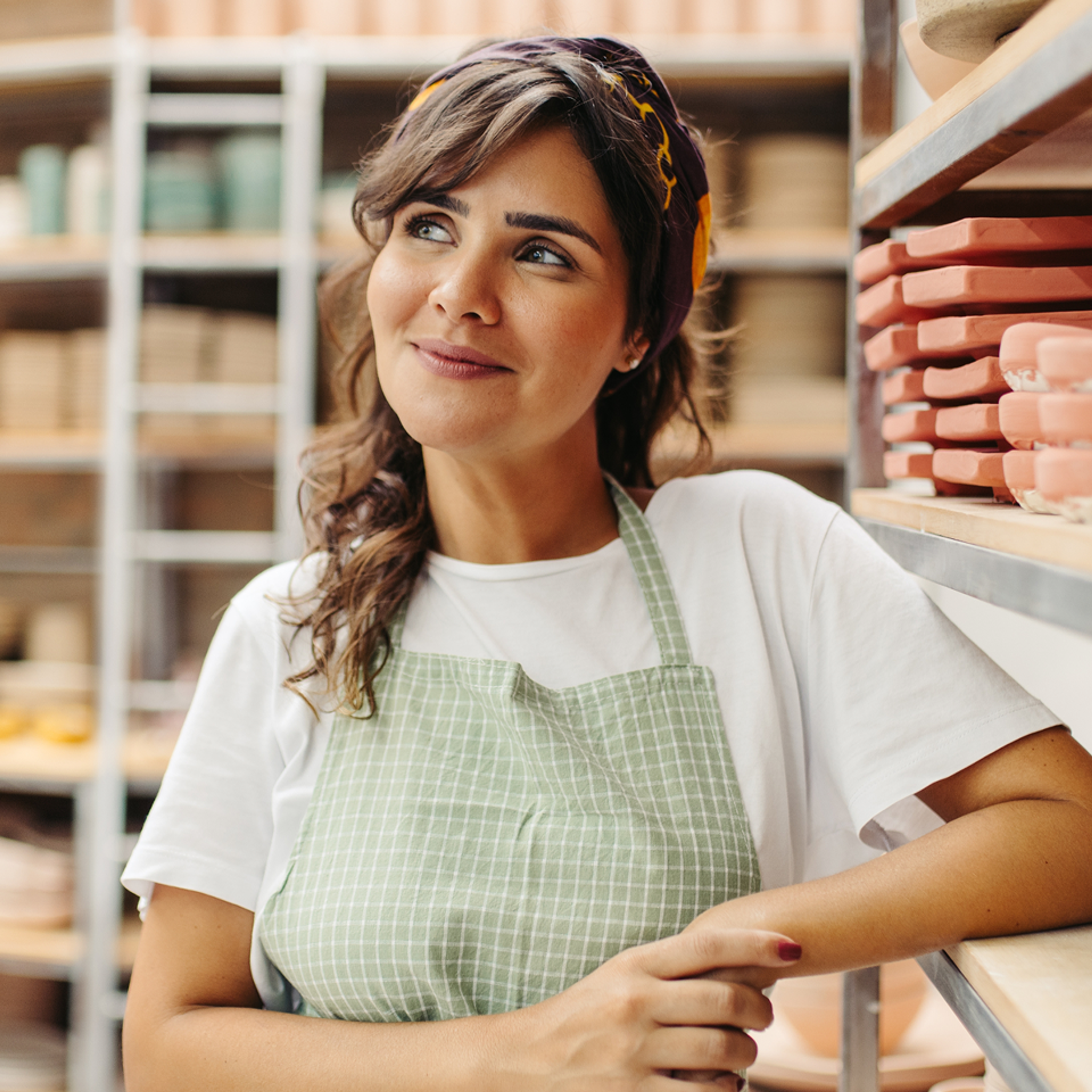 A woman wearing a green checkered apron and a headscarf stands smiling in a pottery studio, surrounded by shelves filled with ceramic pieces.