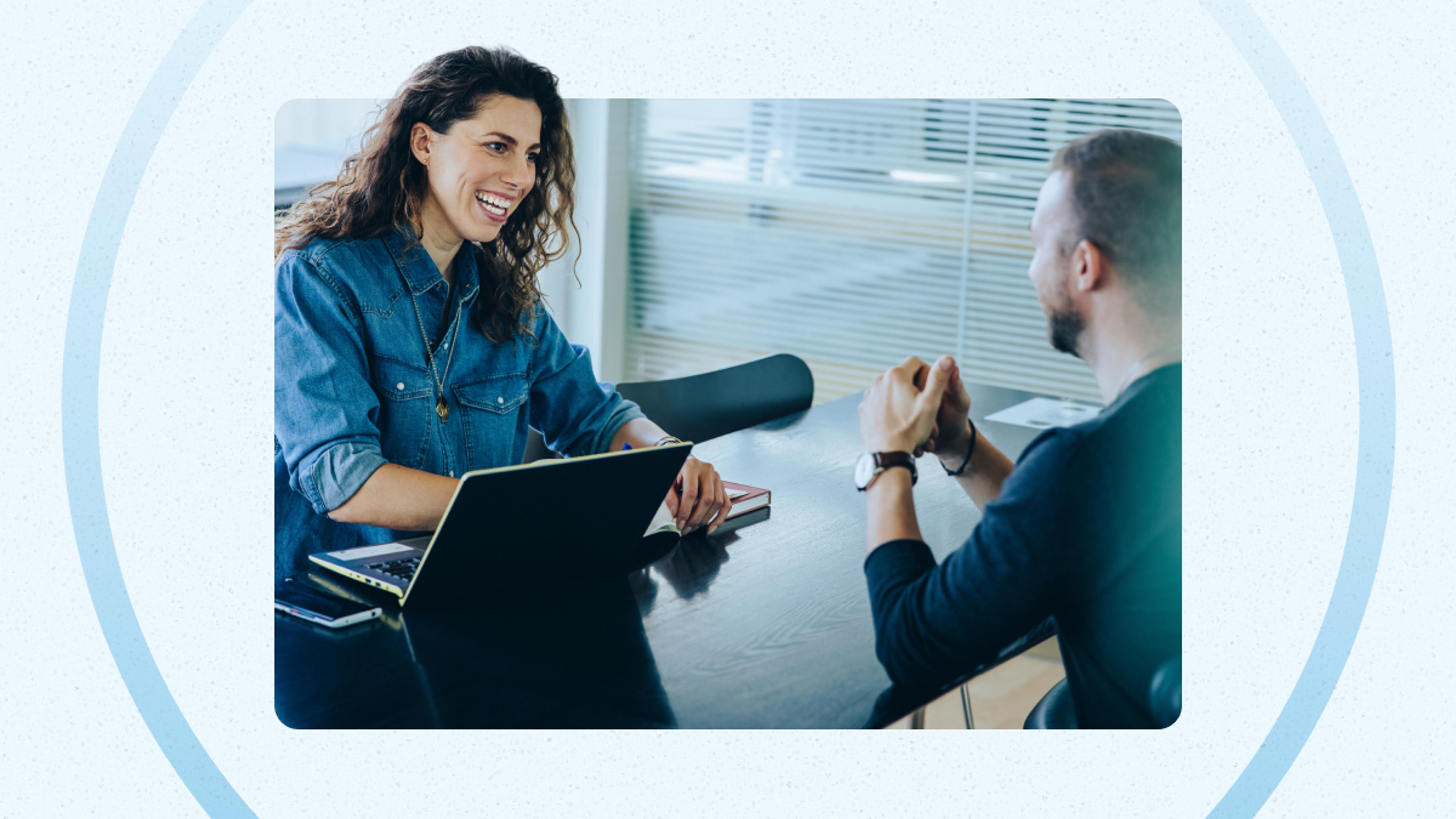 Two people sit across from each other at a table in an office, one with a laptop open, appearing engaged in a conversation.