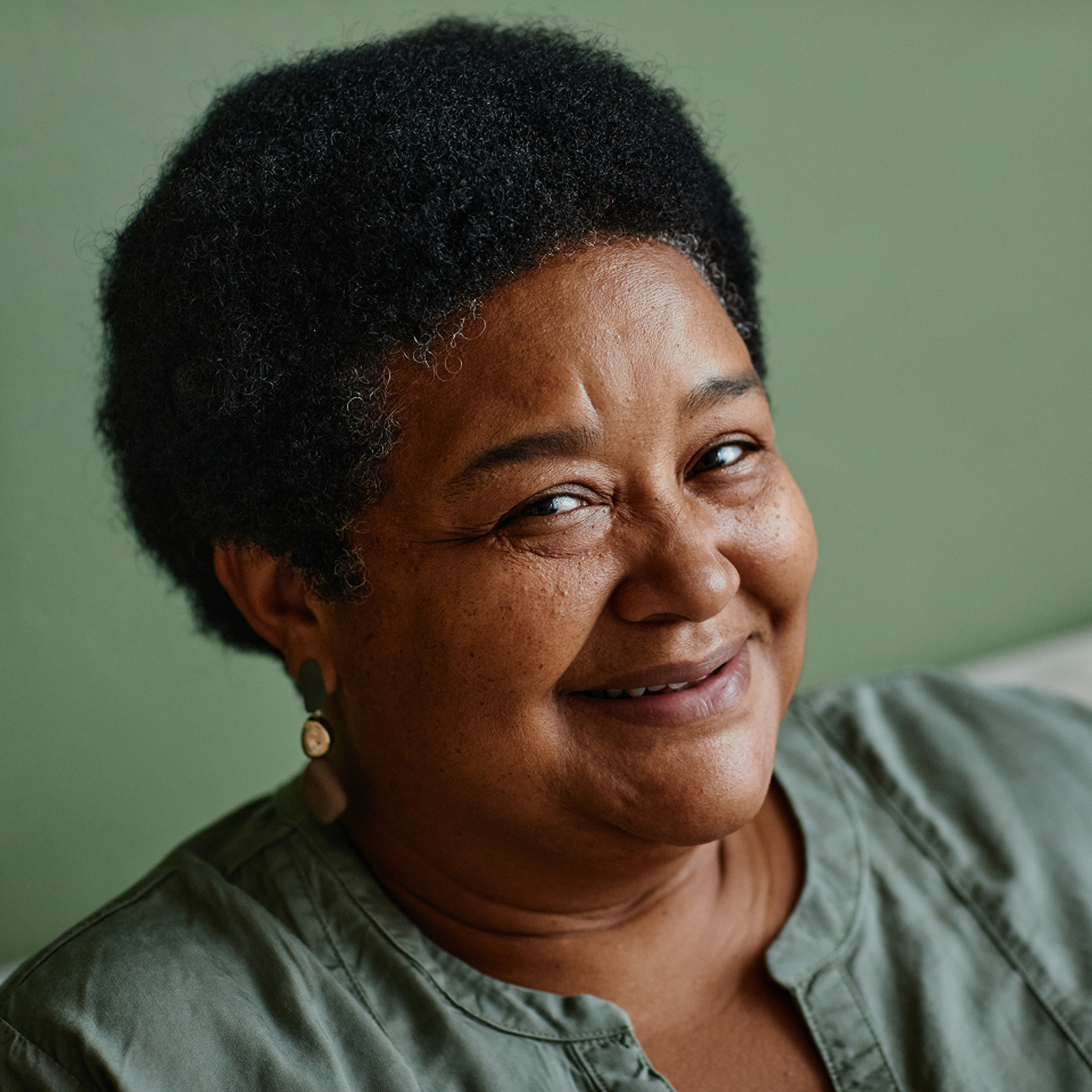 A woman with short curly hair and earrings is smiling slightly, wearing a green top, against a plain green background.