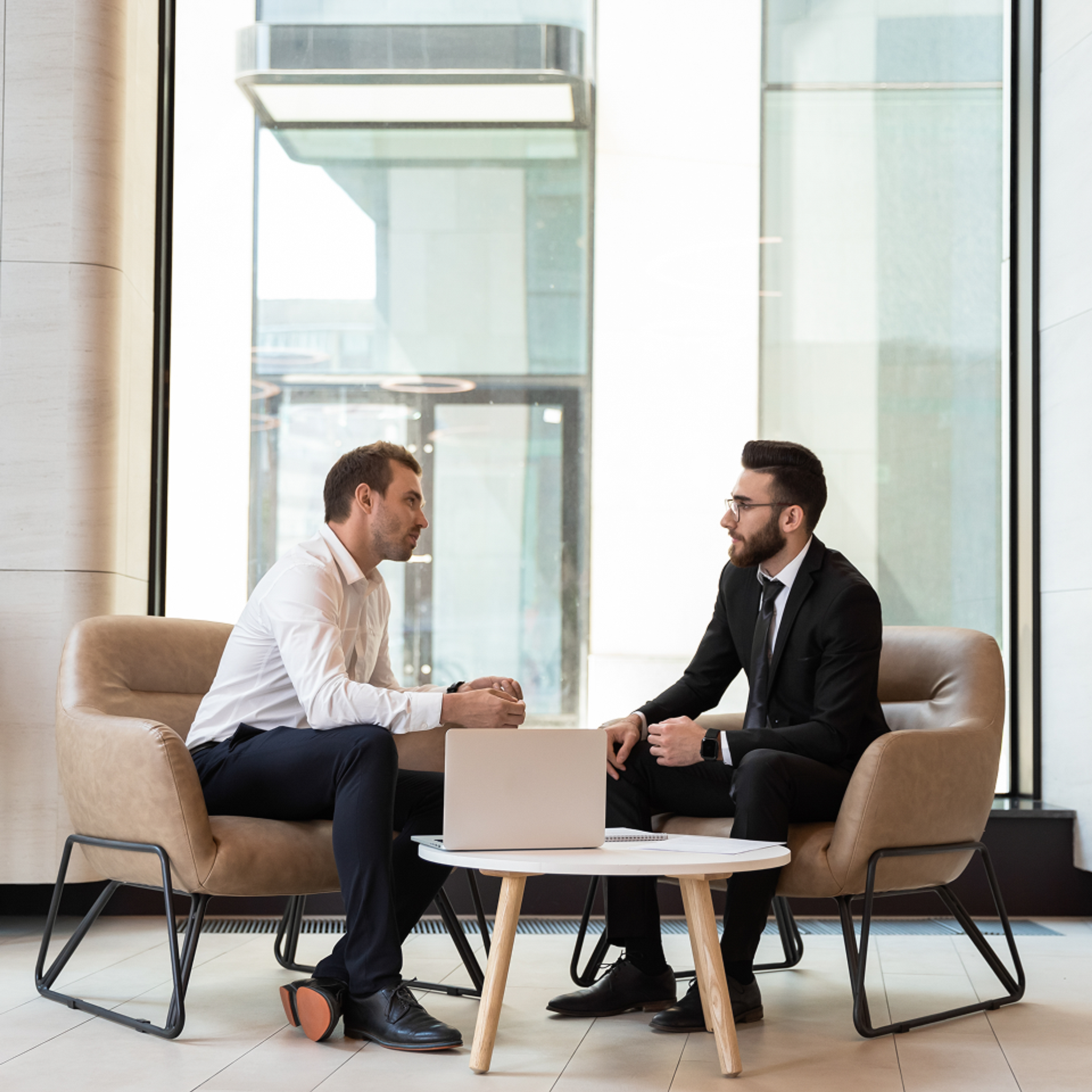 Two men in professional dress seated and discussing with an open computer next to them.
