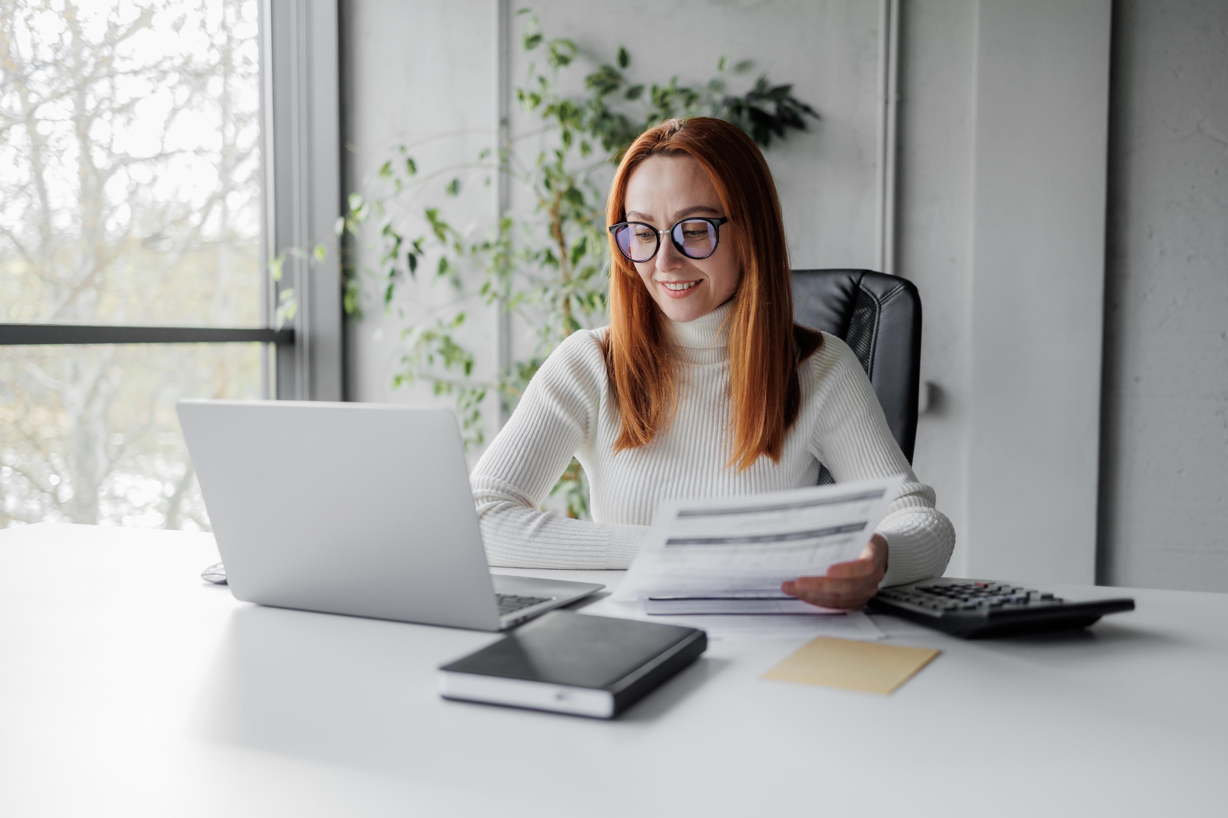 Woman with long red hair and glasses sits at a desk, looking at papers and using a laptop, with a calculator and notebook nearby.