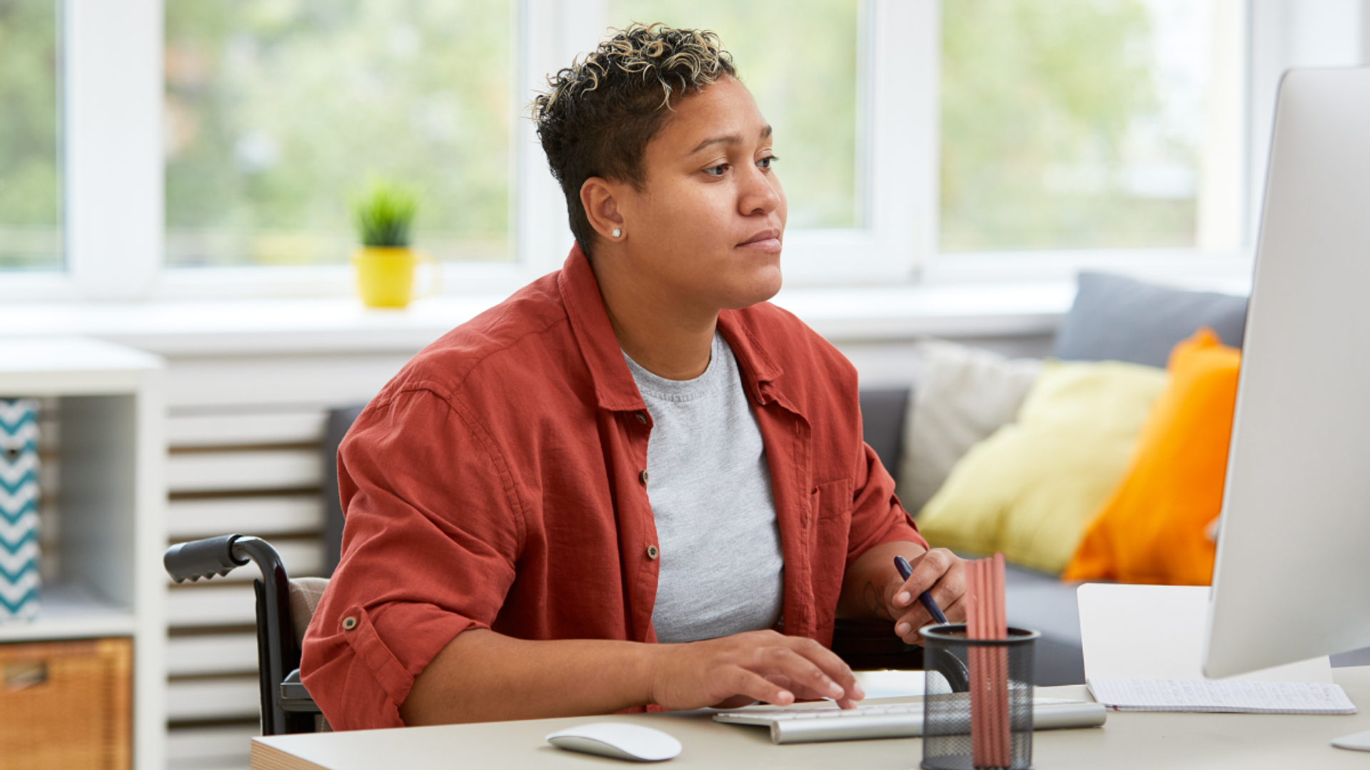 A person sitting in a wheelchair works at a desk, looking at a computer monitor while typing and holding a pen. The background shows a bright, modern living space with large windows and colorful pillows.