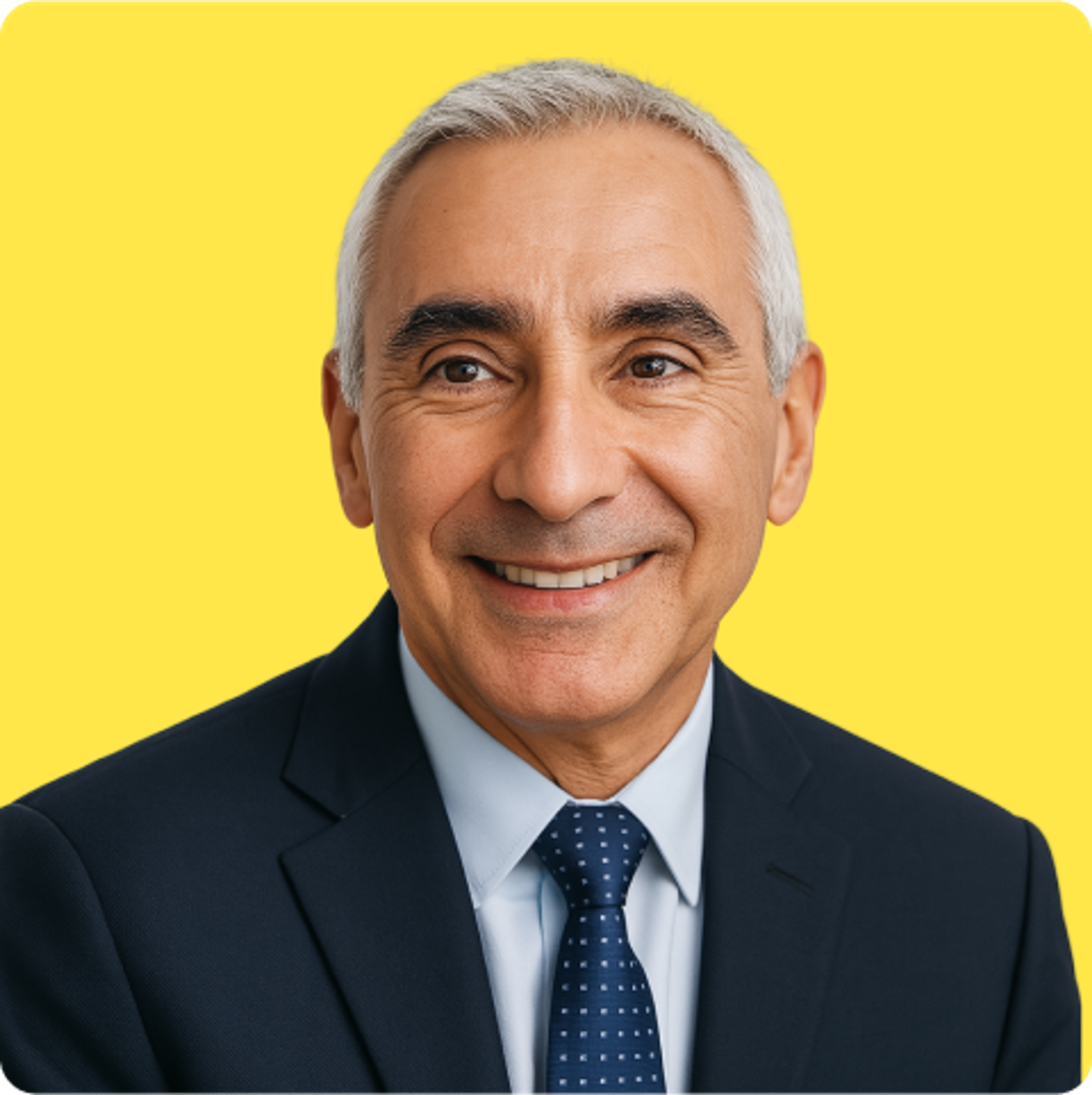 Smiling older man with short gray hair wearing a dark suit, light blue shirt, and navy polka dot tie, posing against a plain yellow background.