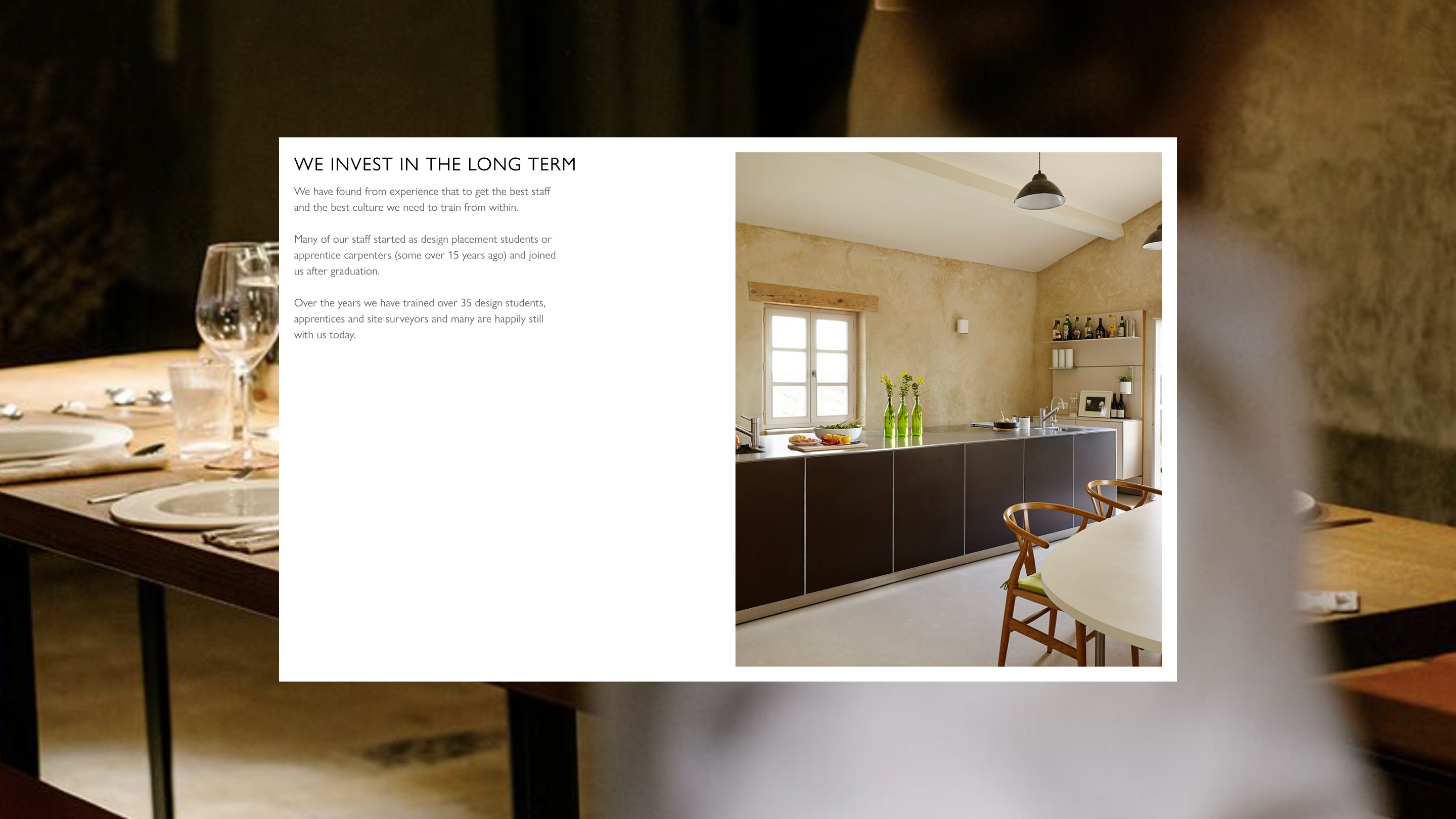 Kitchen Architecture careers page section titled "We Invest In The Long Term," with body copy describing how the company trains staff from within and has trained over 35 design students, apprentices and site surveyors, set alongside a photograph of a rustic plaster-walled kitchen featuring dark cabinetry, a stainless steel worktop, wishbone chairs and a pendant light.