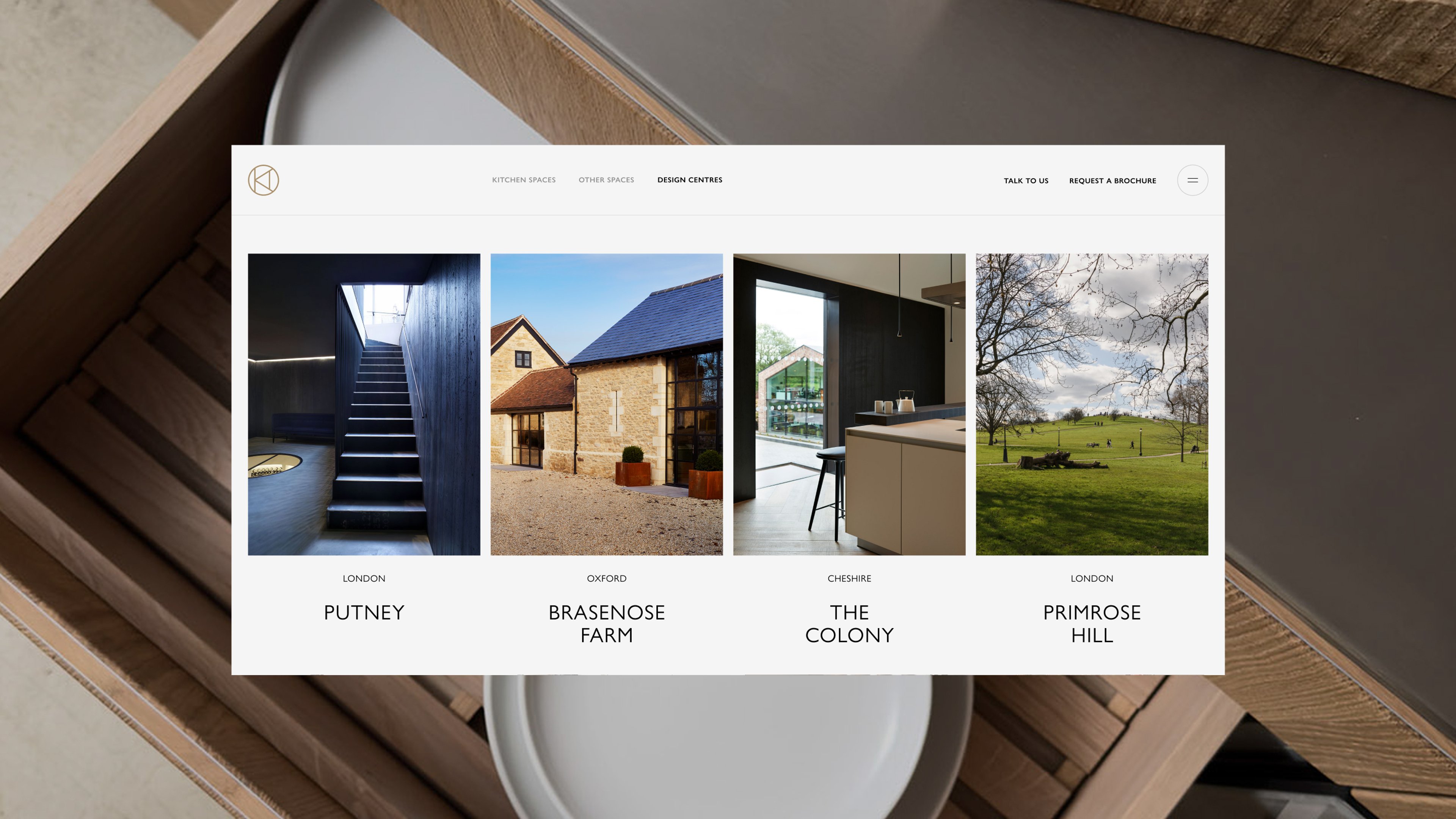 Kitchen Architecture's Design Centres web page set against a top-down photograph of an open wooden drawer, displaying a four-column grid of locations: Putney (London) showing a dark timber staircase, Brasenose Farm (Oxford) showing a converted stone barn, The Colony (Cheshire) showing a contemporary kitchen interior, and Primrose Hill (London) showing a parkland view.