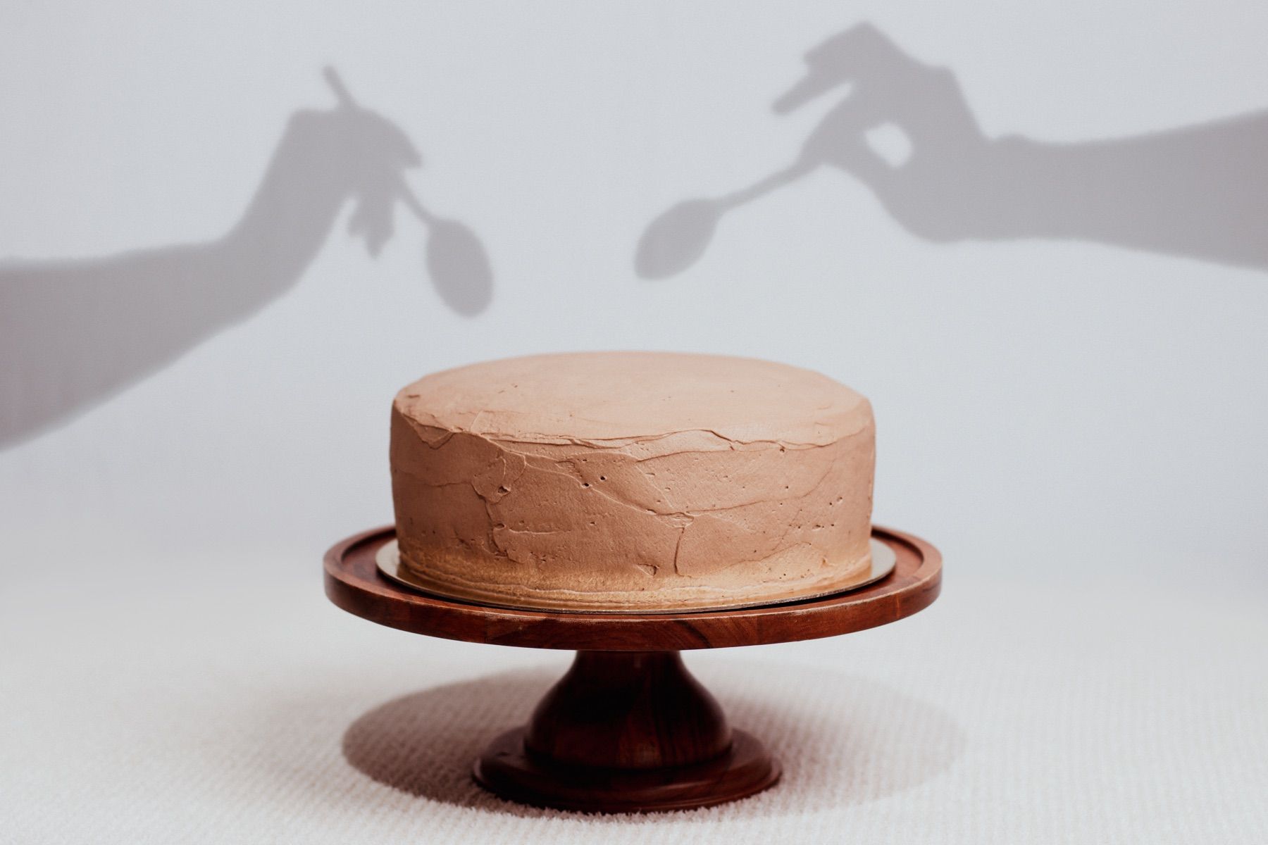 Round cake with smooth icing on a dark wood pedestal stand, with playful shadow-puppet hands reaching toward it