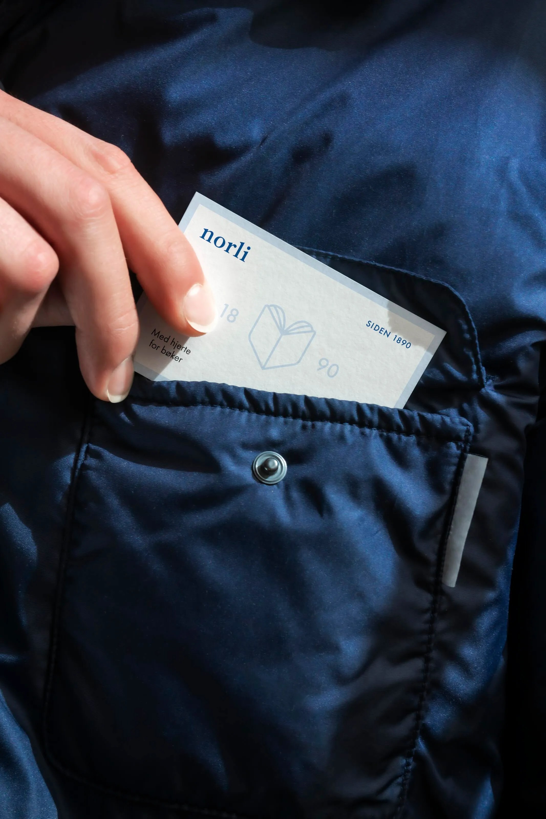 Hand placing a Norli business card with book logo into a navy blue pocket.