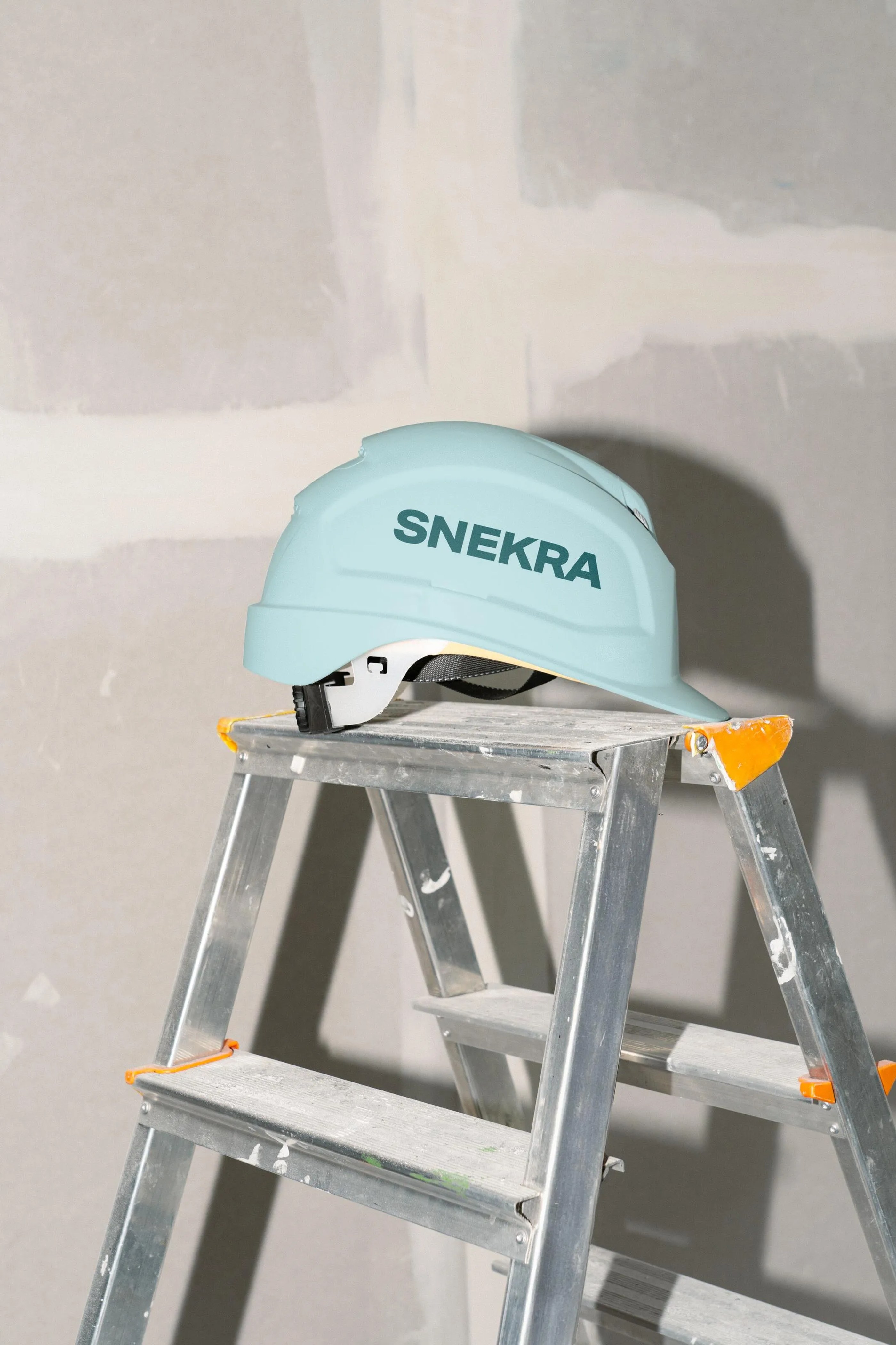 Light blue construction helmet with the word SNEKRA placed on top of a metal step ladder against a partially finished drywall background.