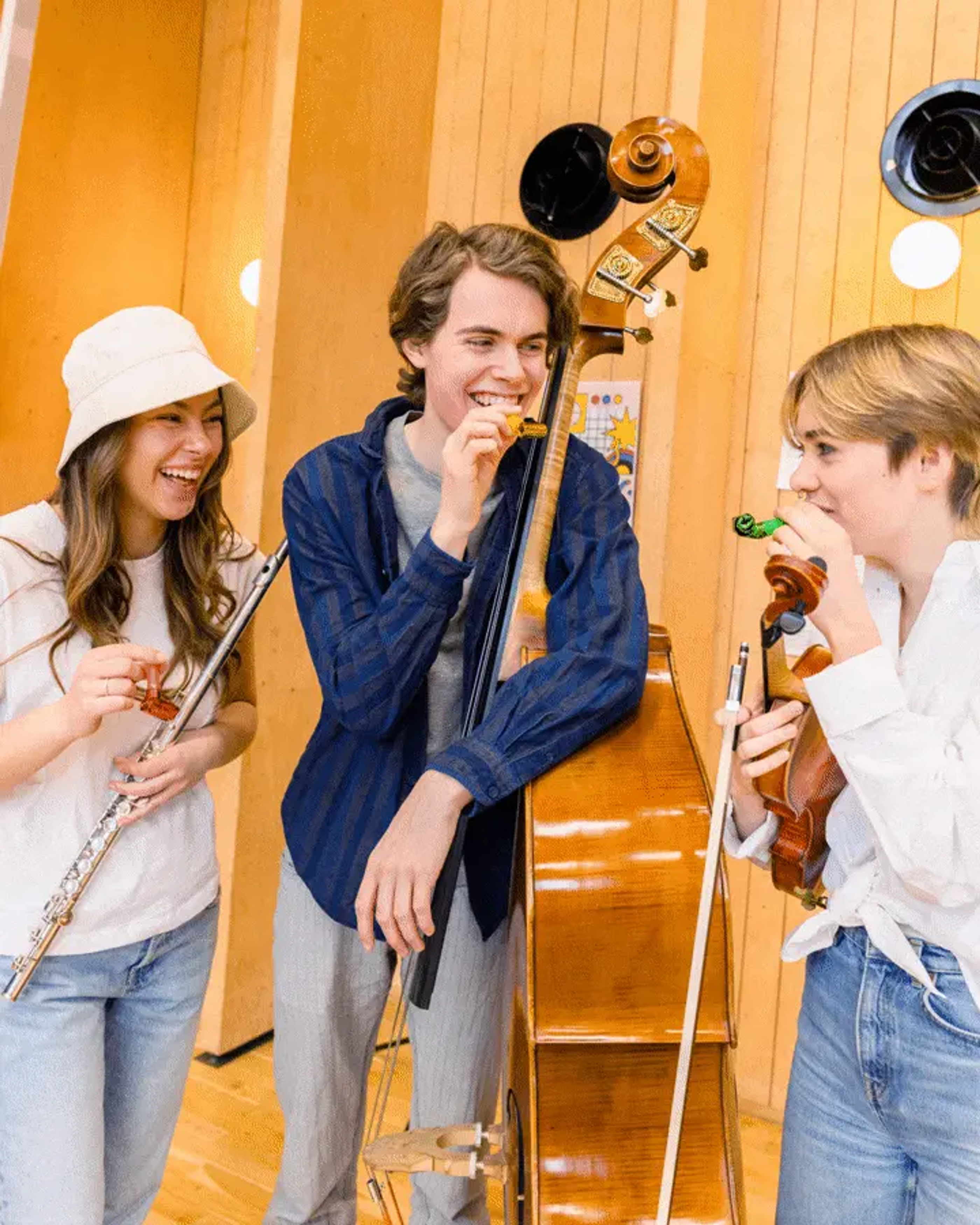 Teen musicians laughing together in a wooden rehearsal room, holding flute, double bass and violin, capturing the playful community feeling of Orkesteråret ensembles.