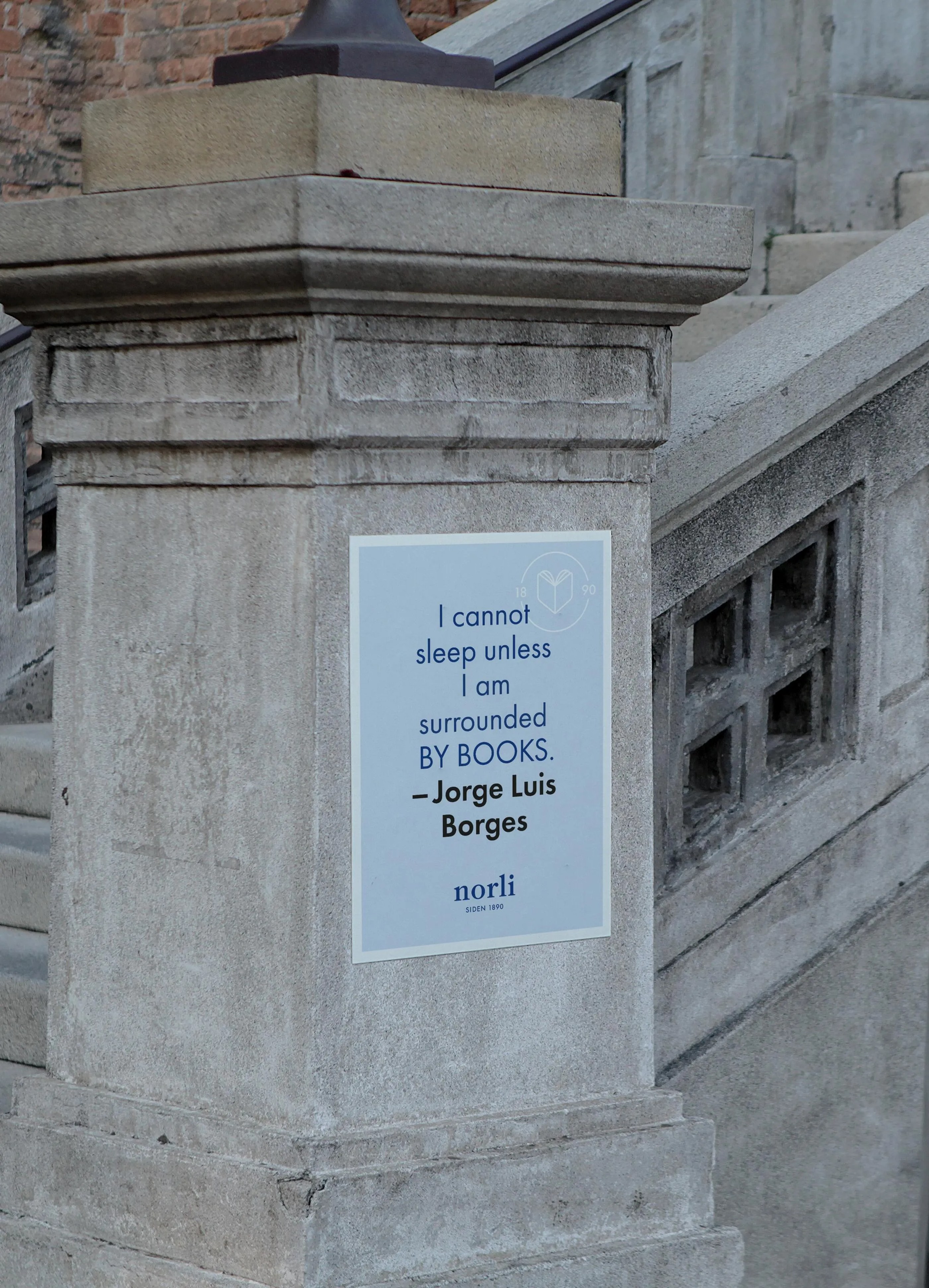 Blue poster on a stone pillar with a quote by Jorge Luis Borges reading, 'I cannot sleep unless I am surrounded by books.'