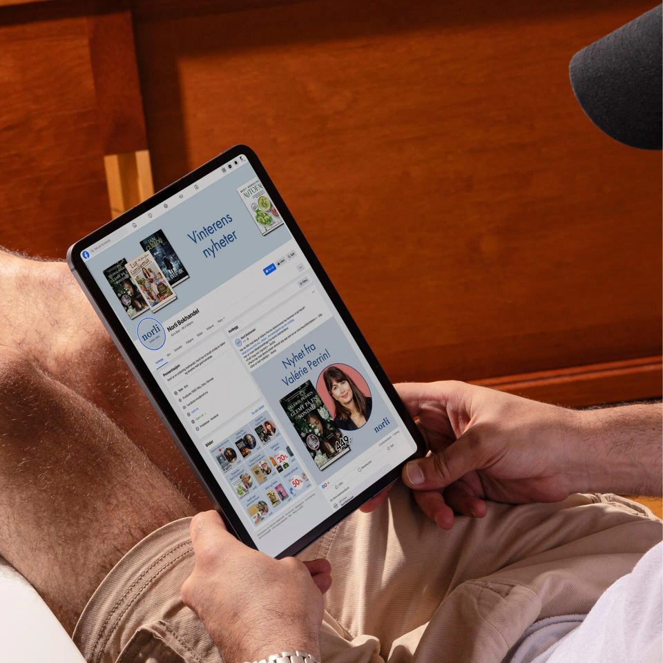 Person holding a tablet displaying Norli Bokhandel Facebook page featuring winter book news and a photo of author Valerie Perrin.