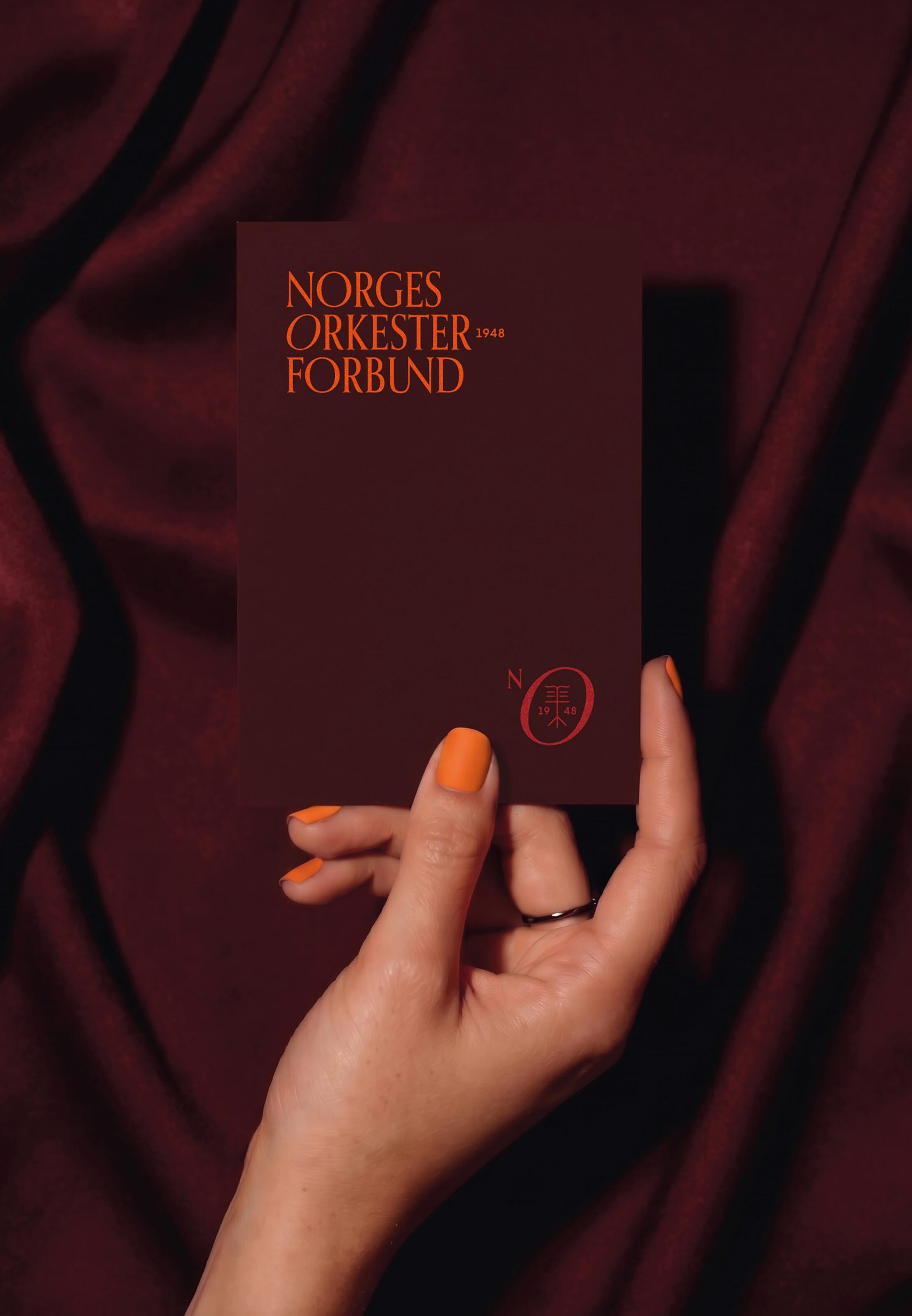 Hand with orange nails holding a burgundy Norges Orkesterforbund card with the orange wordmark and secondary symbol.