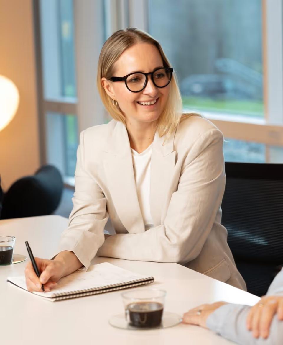 Aimee smiling. She has blonde hair and glasses writing in a notebook at a white table with coffee cups.