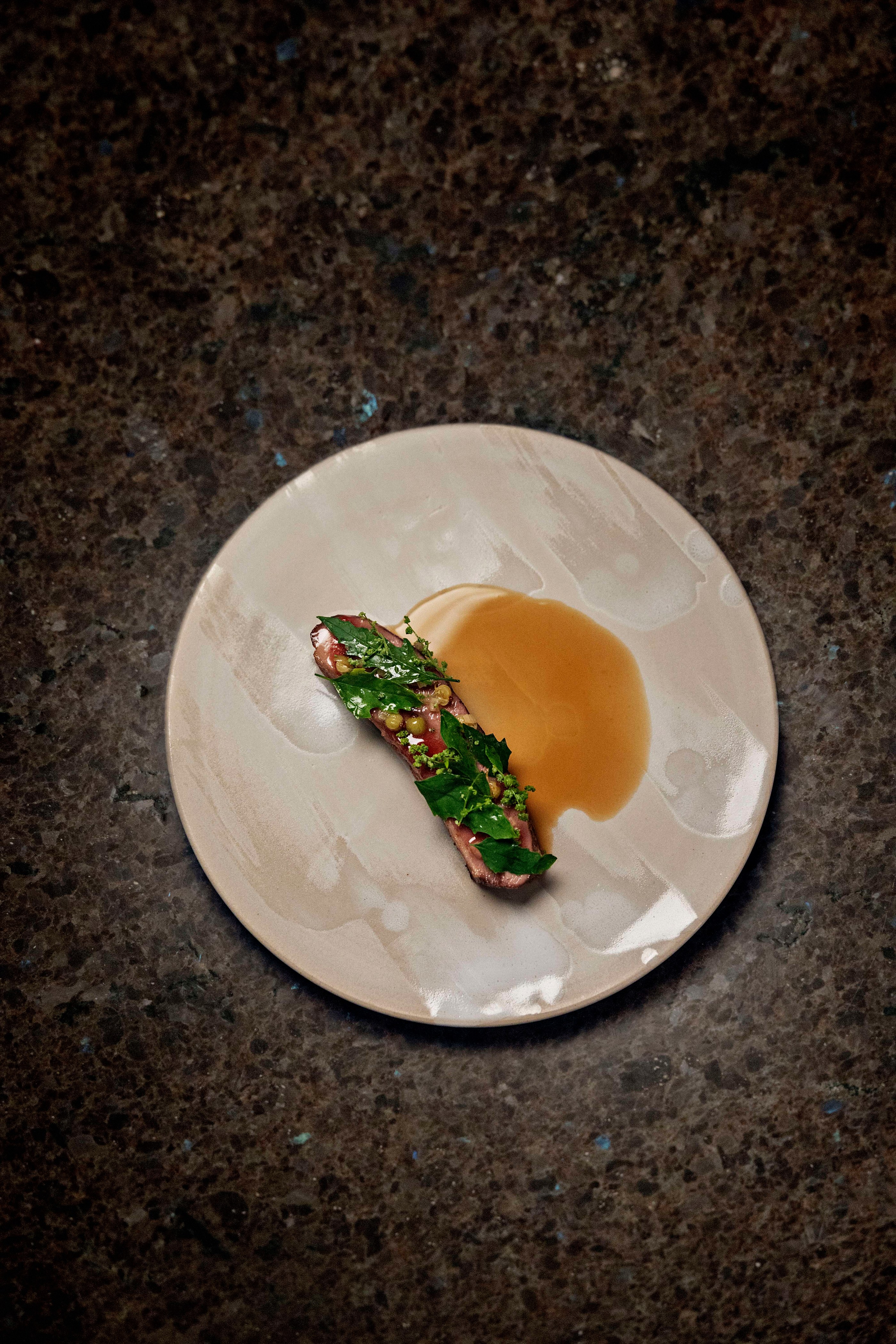 Minimalist gourmet dish with a piece of meat garnished with green herbs and a light brown sauce on a beige plate.