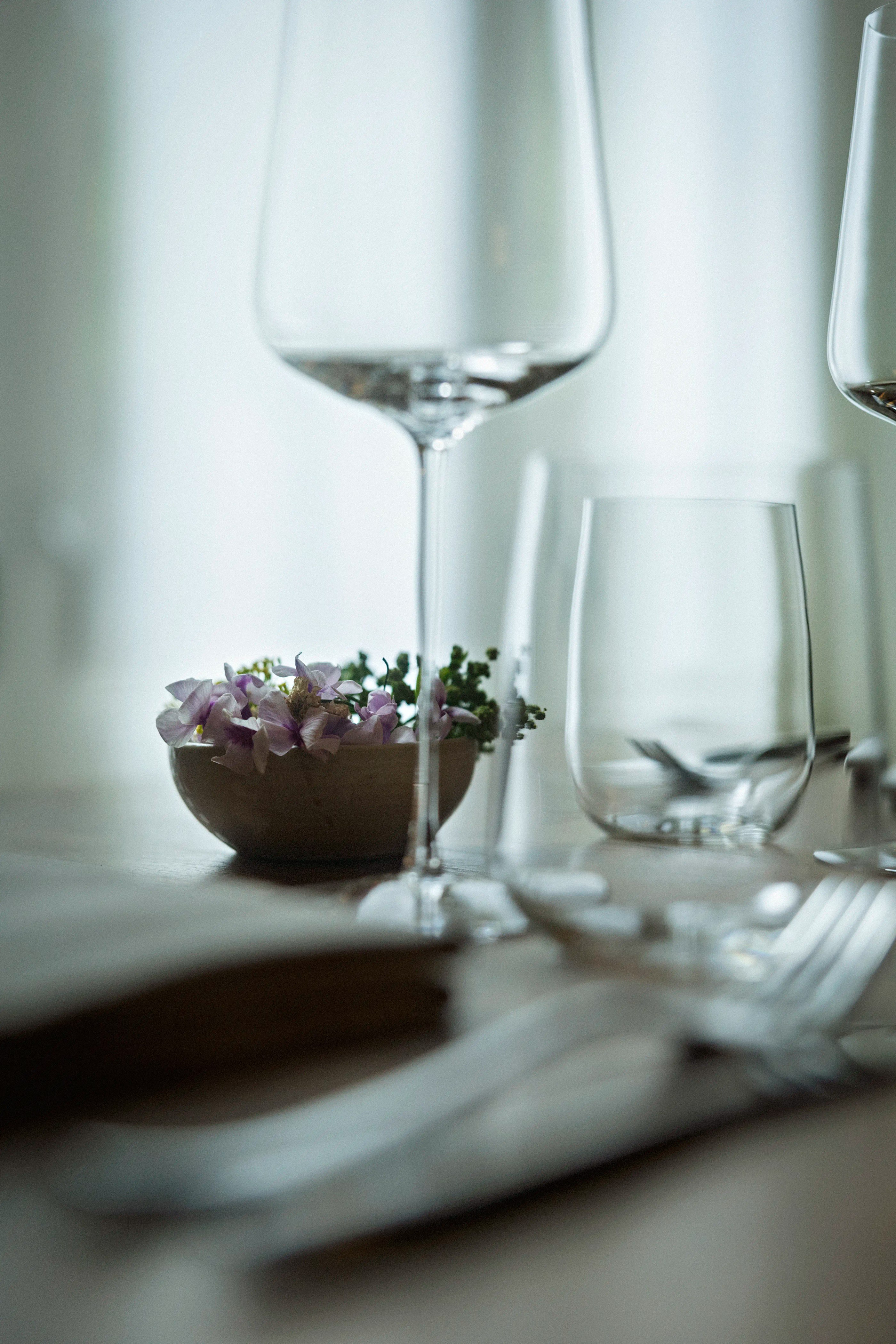 Close-up of a dining table with empty wine and water glasses, silverware, and a small bowl of purple flowers and greenery.