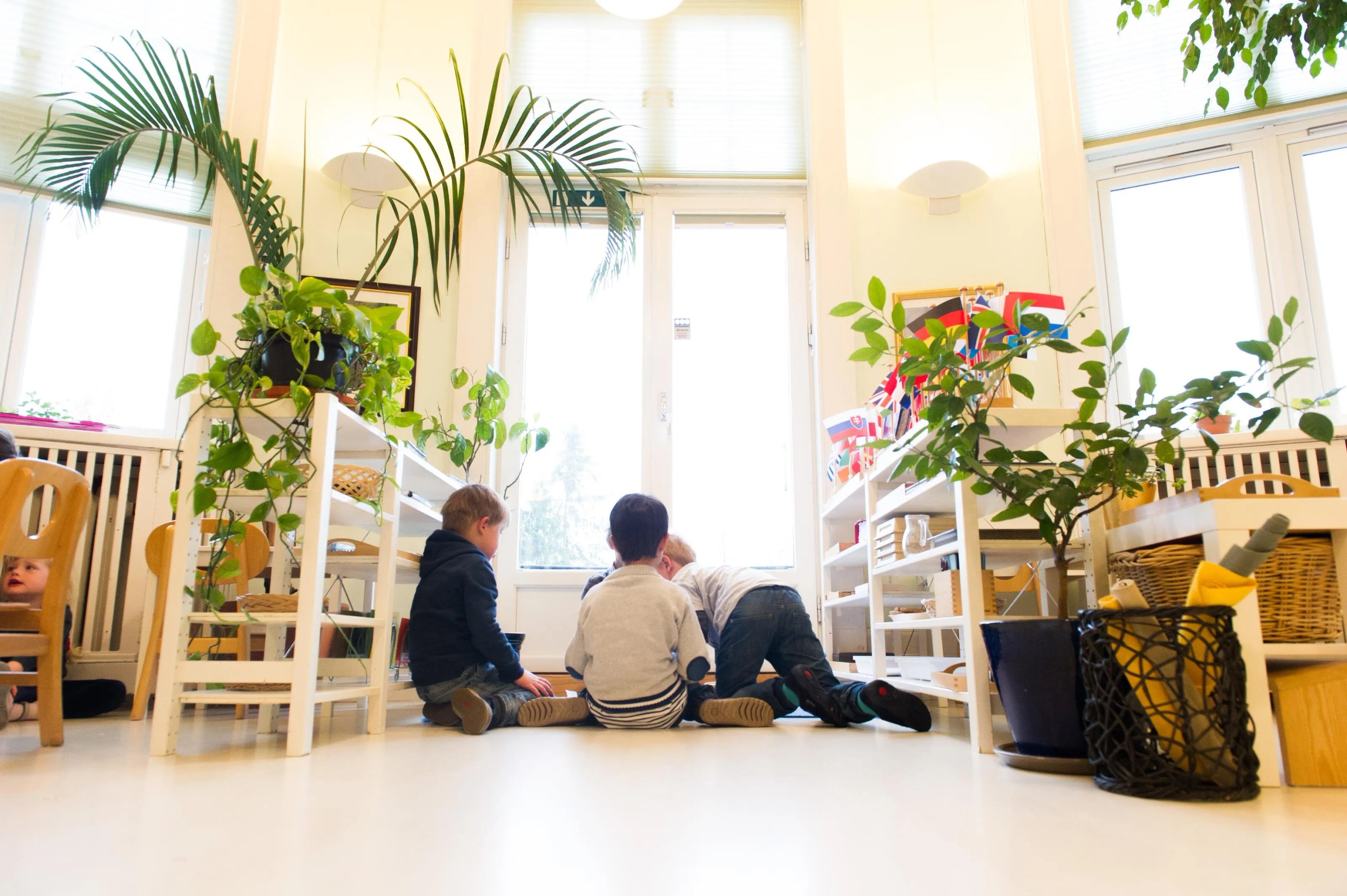Three children sitting on the floor in a bright classroom surrounded by plants and shelves, with one child partially visible under a chair.