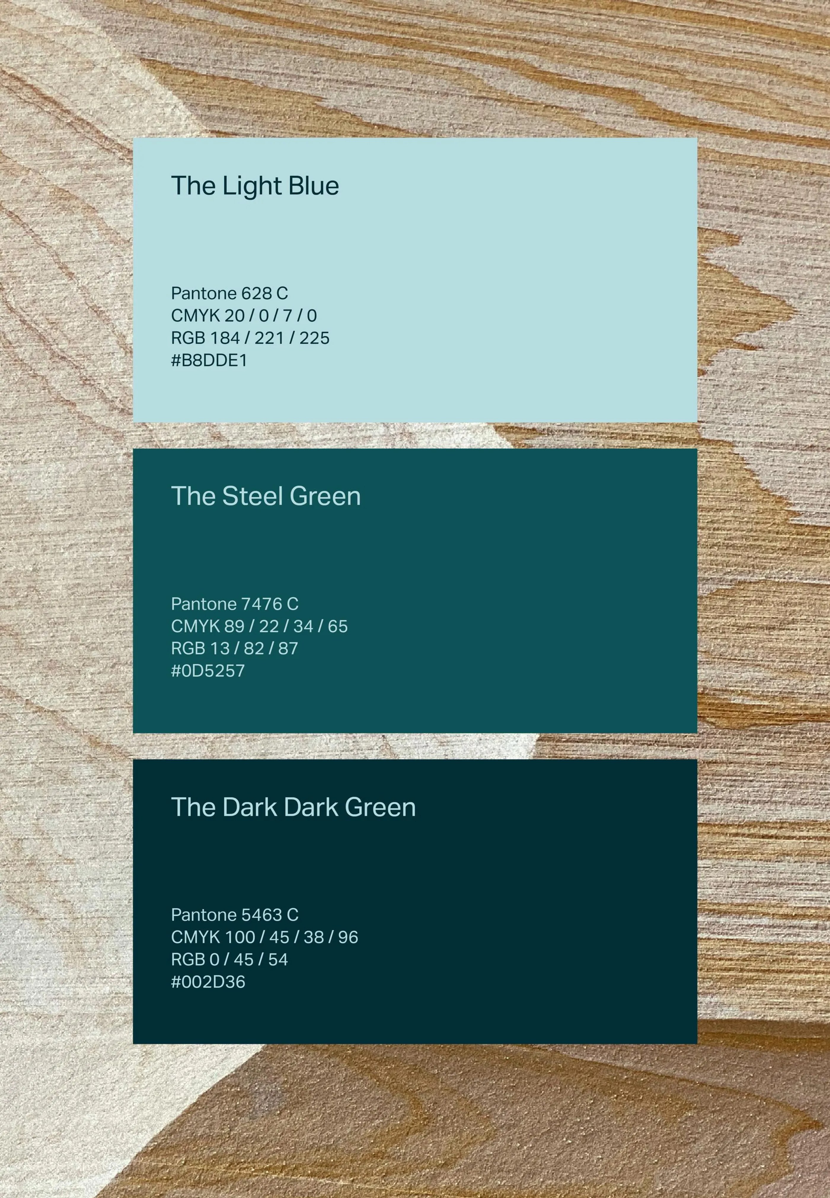 Three color swatches on a wooden surface showing The Light Blue, The Steel Green, and The Dark Dark Green with Pantone, CMYK, RGB, and hex codes.
