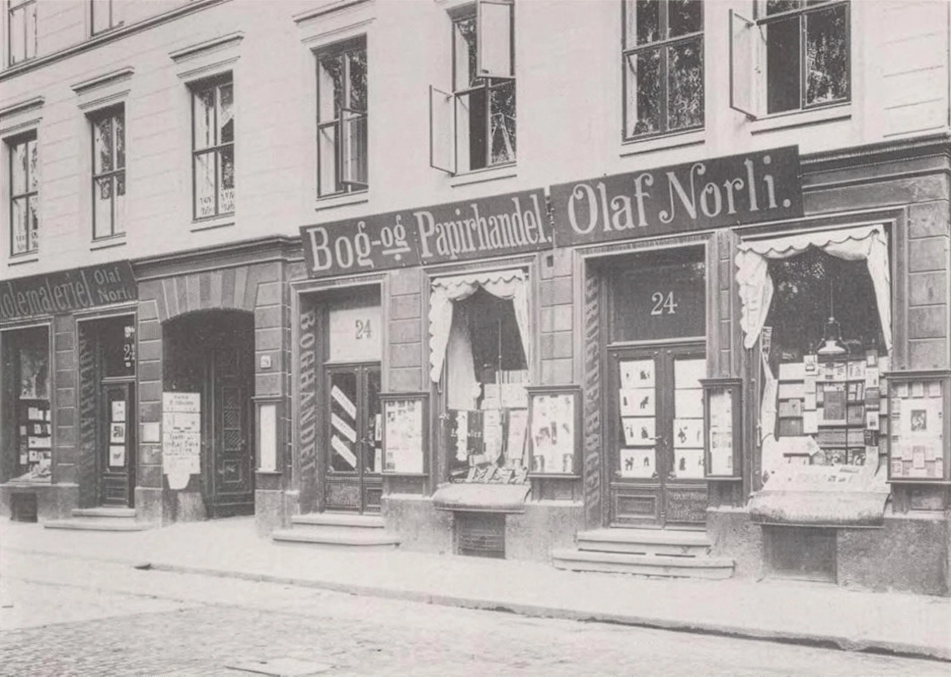 Black and white photo of the first Norli bookstore and stationery shop with signs reading 'Bog-og Papirhandel Olaf Norli' and the address number 24 above the doors.