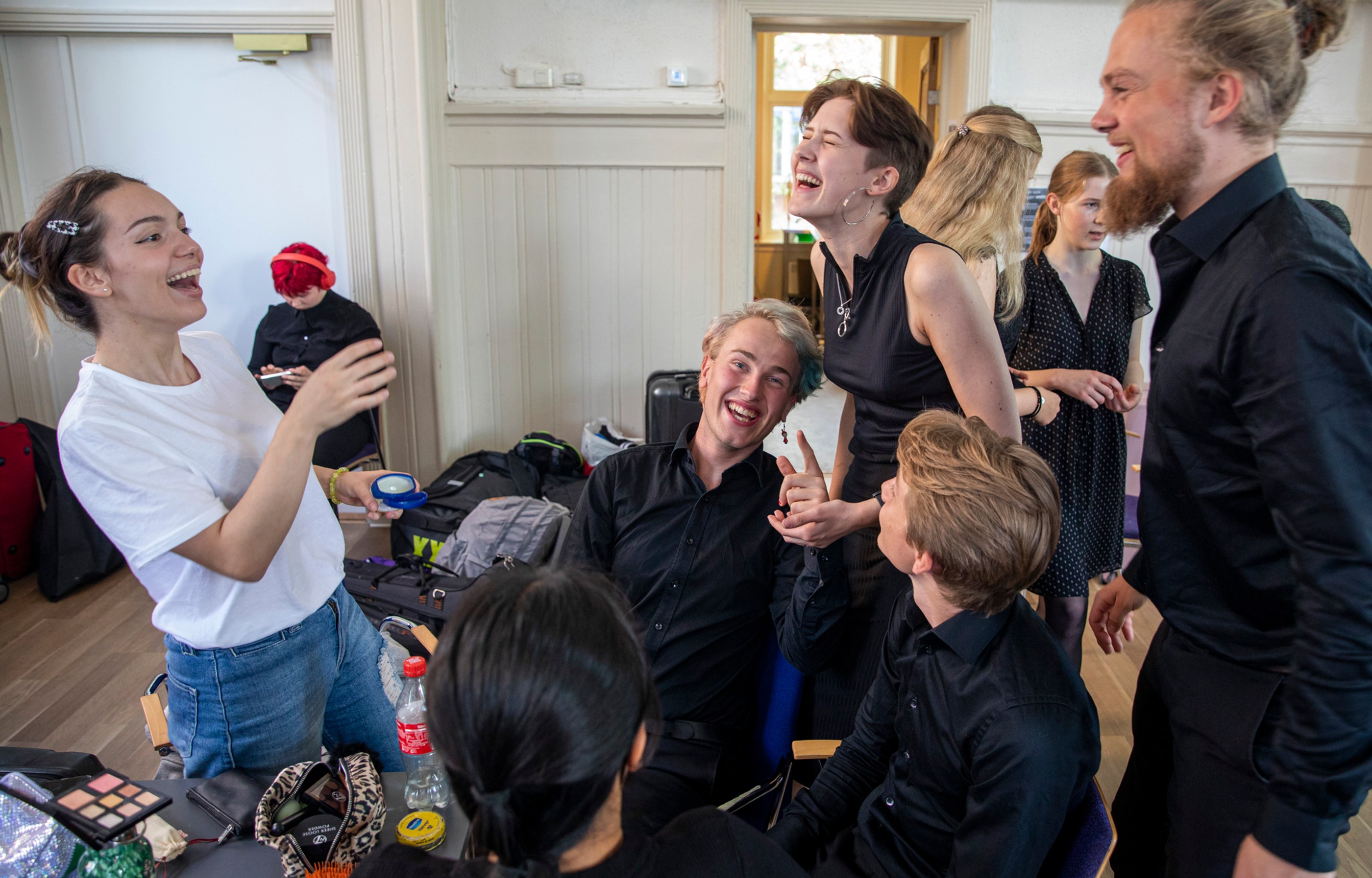 Group of young people laughing and socializing indoors, some sitting and some standing near a table with makeup and a drink bottle.
