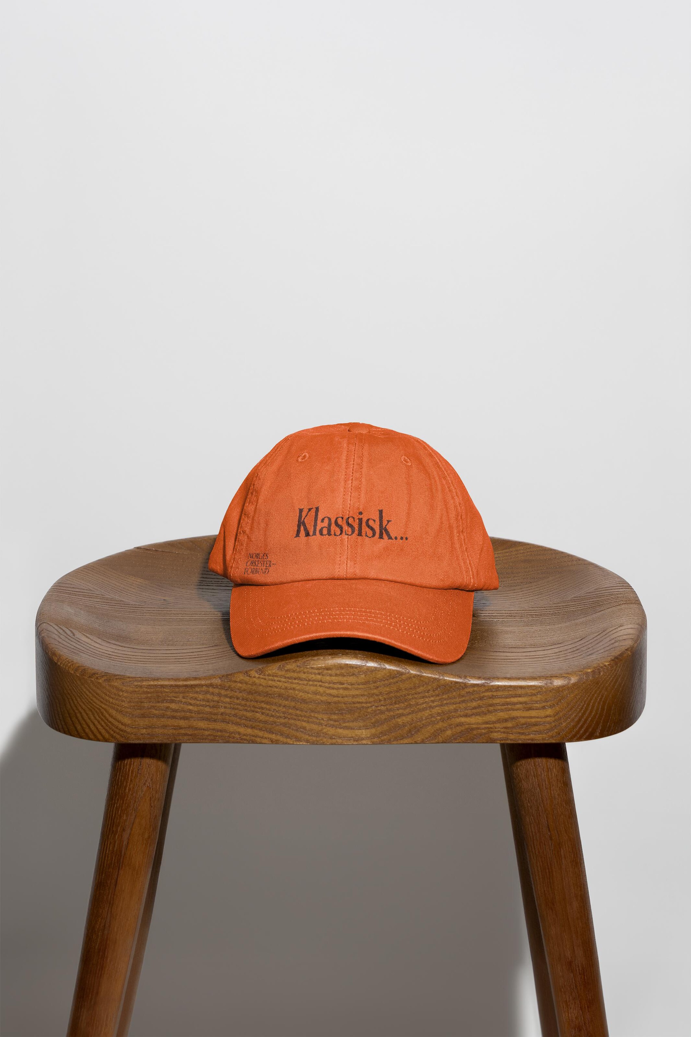 Orange cap embroidered with “Klassisk…” and a small Norges Orkesterforbund wordmark, resting on a wooden stool.