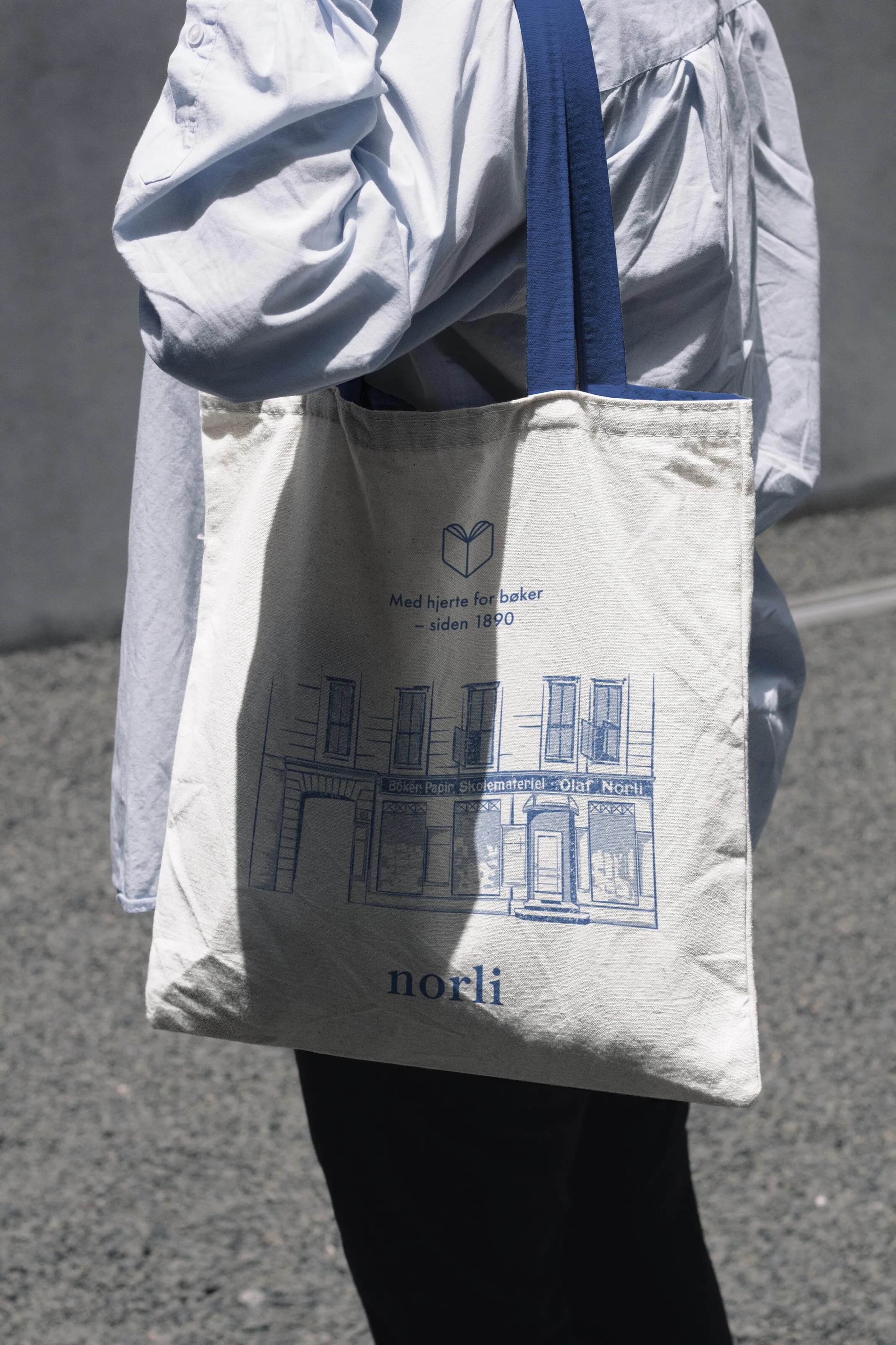 Person wearing a white shirt carrying a cream tote bag with blue handles and a blue illustration of a building and text reading 'Med hjerte for bøker – siden 1890' and 'norli'.