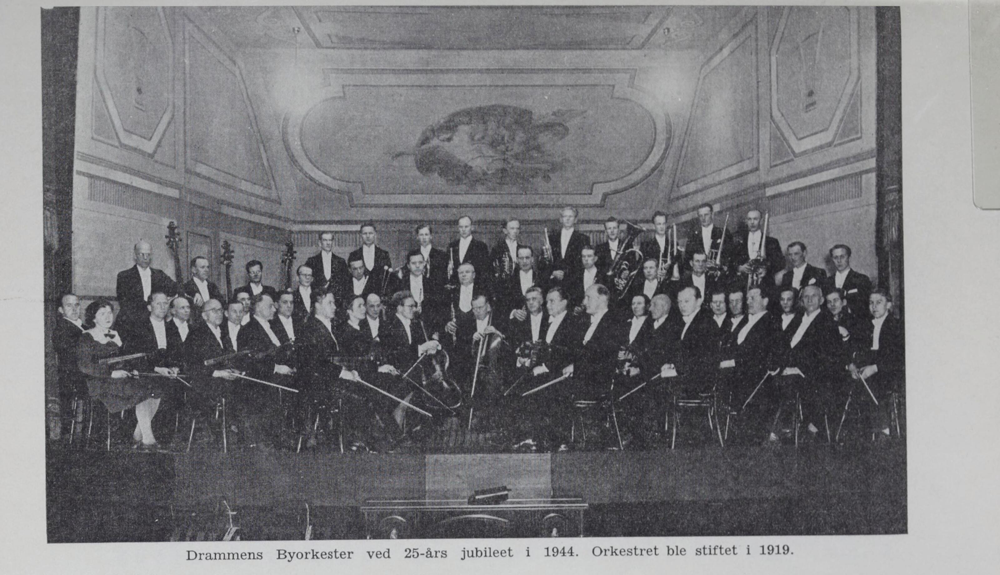 Black-and-white archive group portrait of Drammen Bysorkester 25th anniversary in 1944 with its conductor in a decorated concert hall.