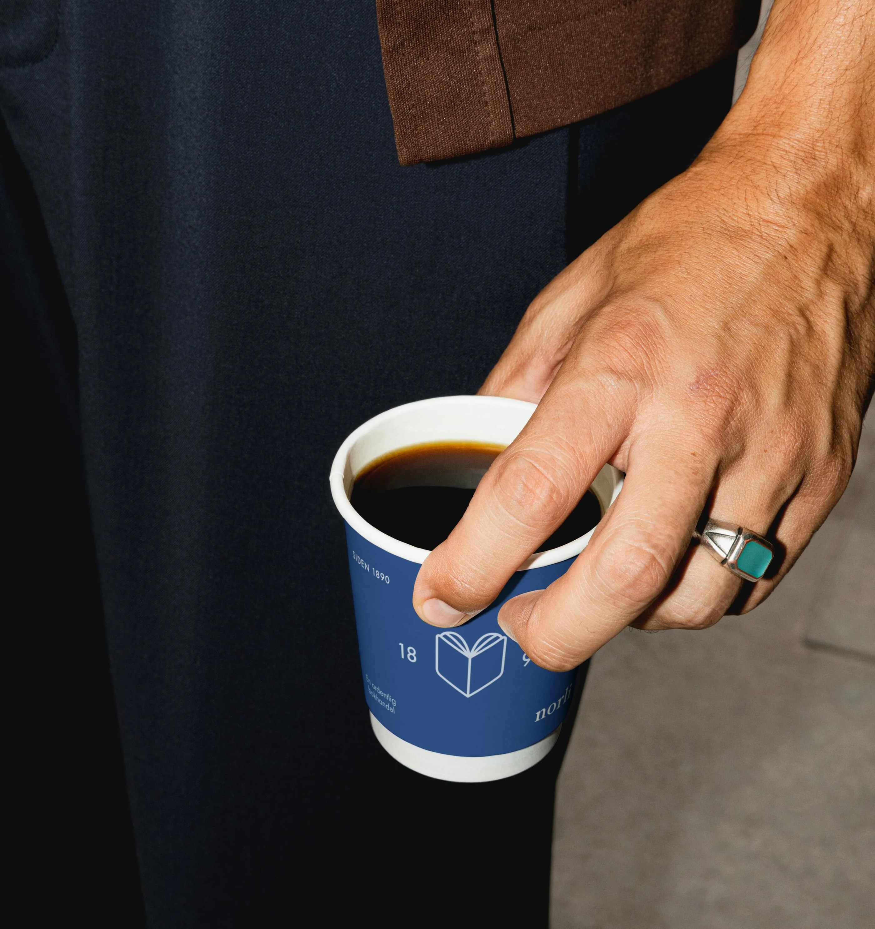 Hand with a silver ring holding a blue paper cup filled with black coffee.