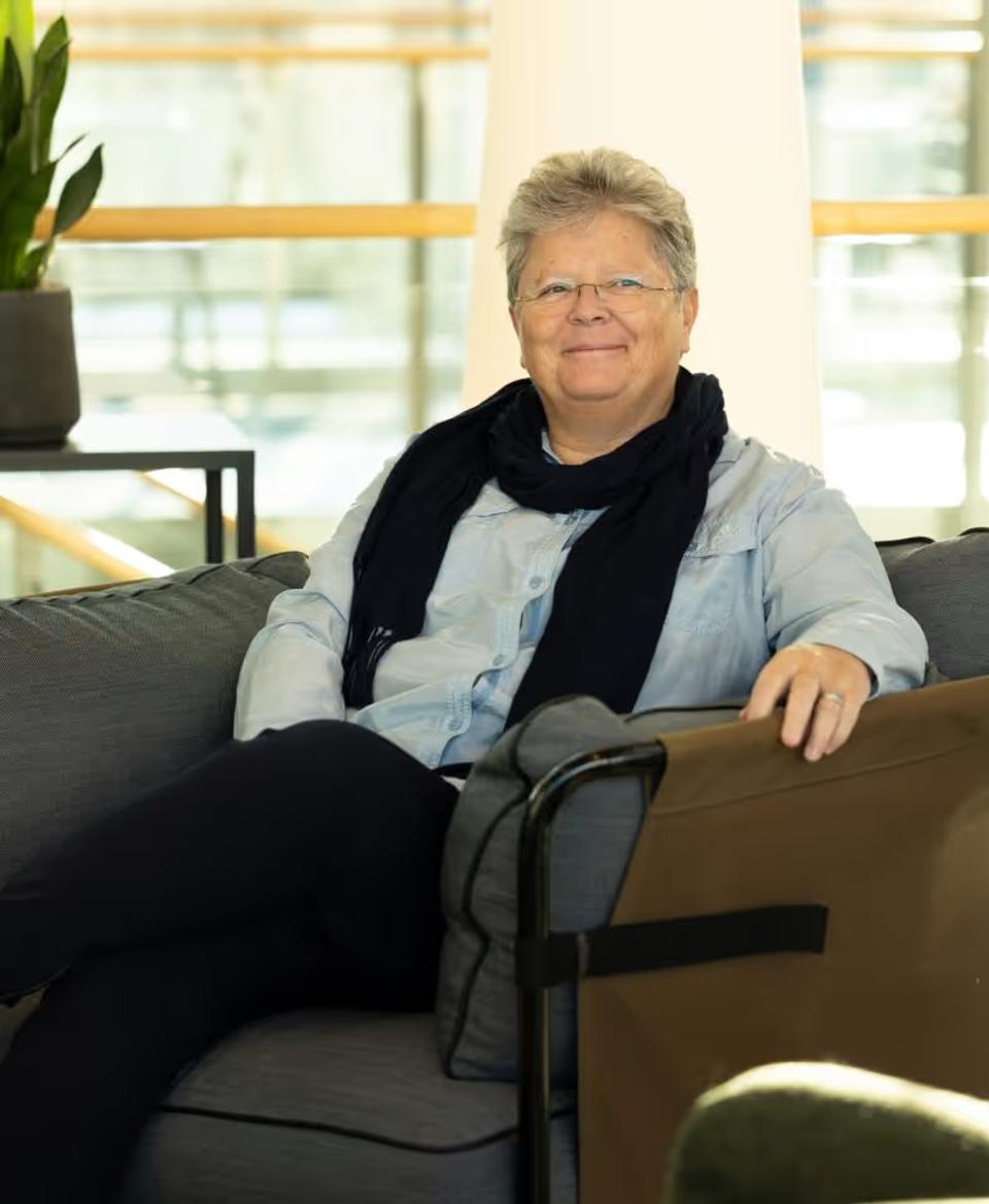 Gerd smiling. She has short hair and glasses sitting on a sofa wearing a light blue shirt and black scarf.