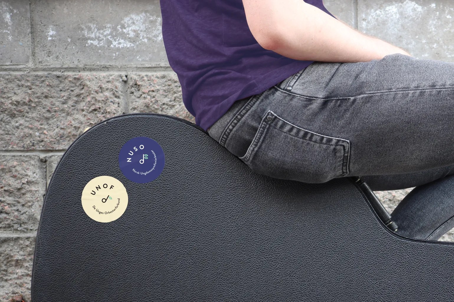 Person sitting on a black instrument case with two round stickers labeled NUSO and UNOF on it against a textured stone wall background.