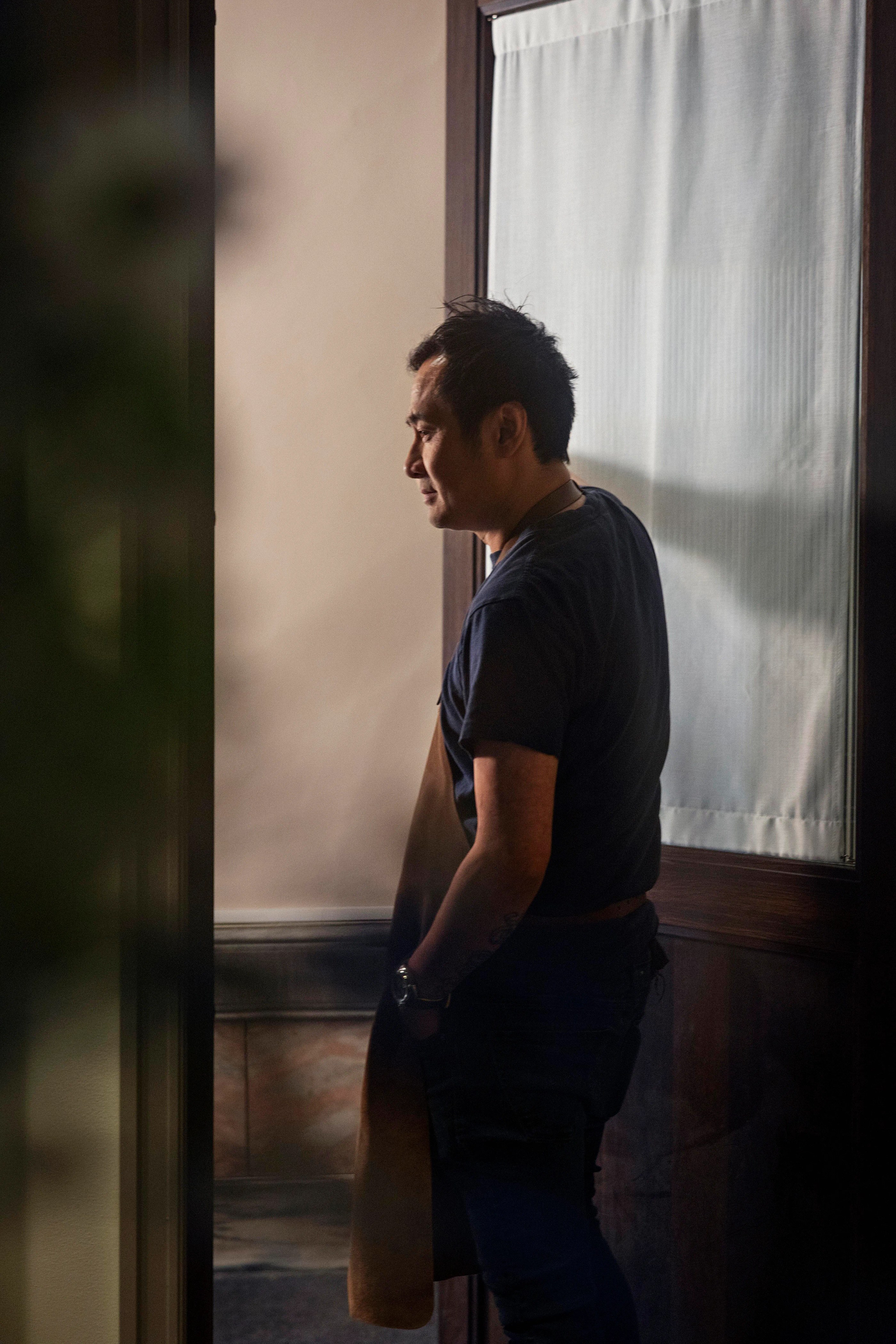 Björn Svensson in a dark shirt wearing an apron stands indoors near a door with a window covered by a curtain.