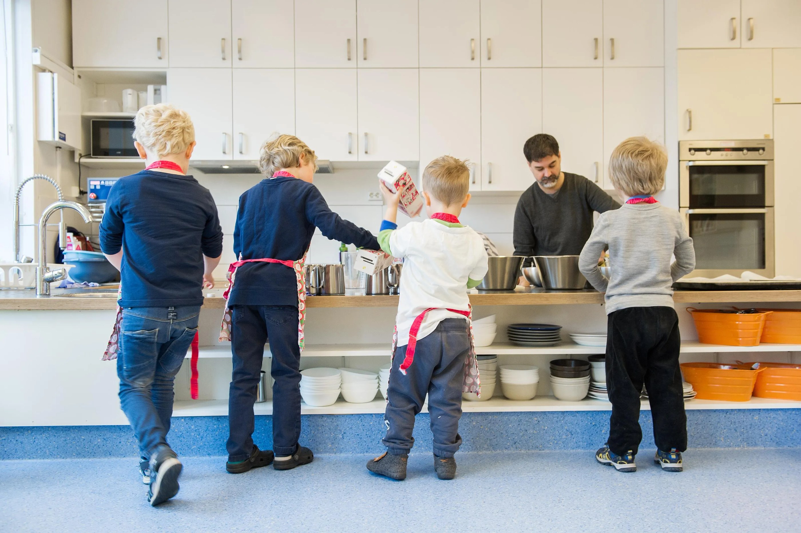 Four children wearing aprons stand at a kitchen counter with a man preparing food behind the counter.