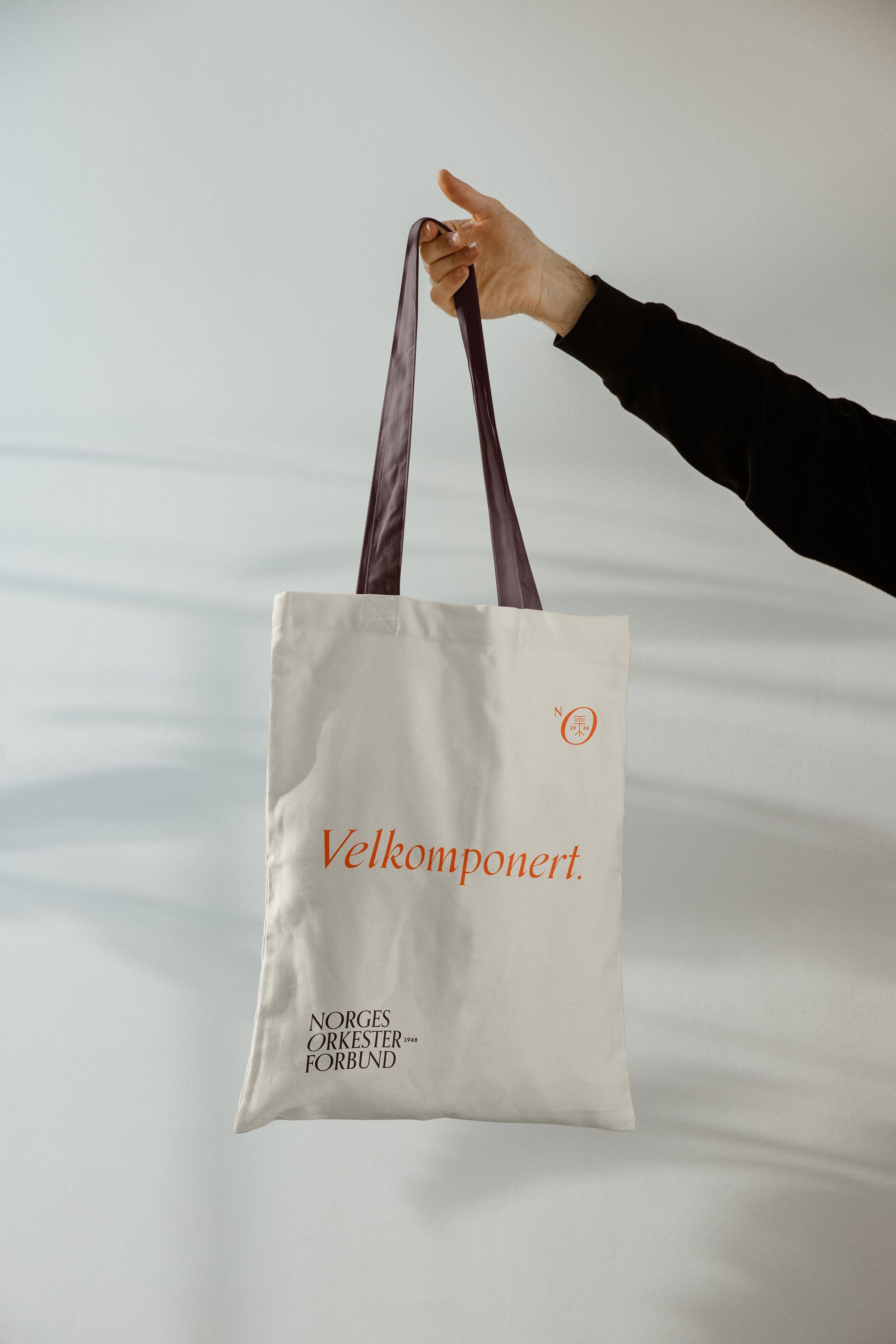 Cream tote bag with burgundy handles, the wordmark, orange secondary symbol and the word “Velkomponert”.