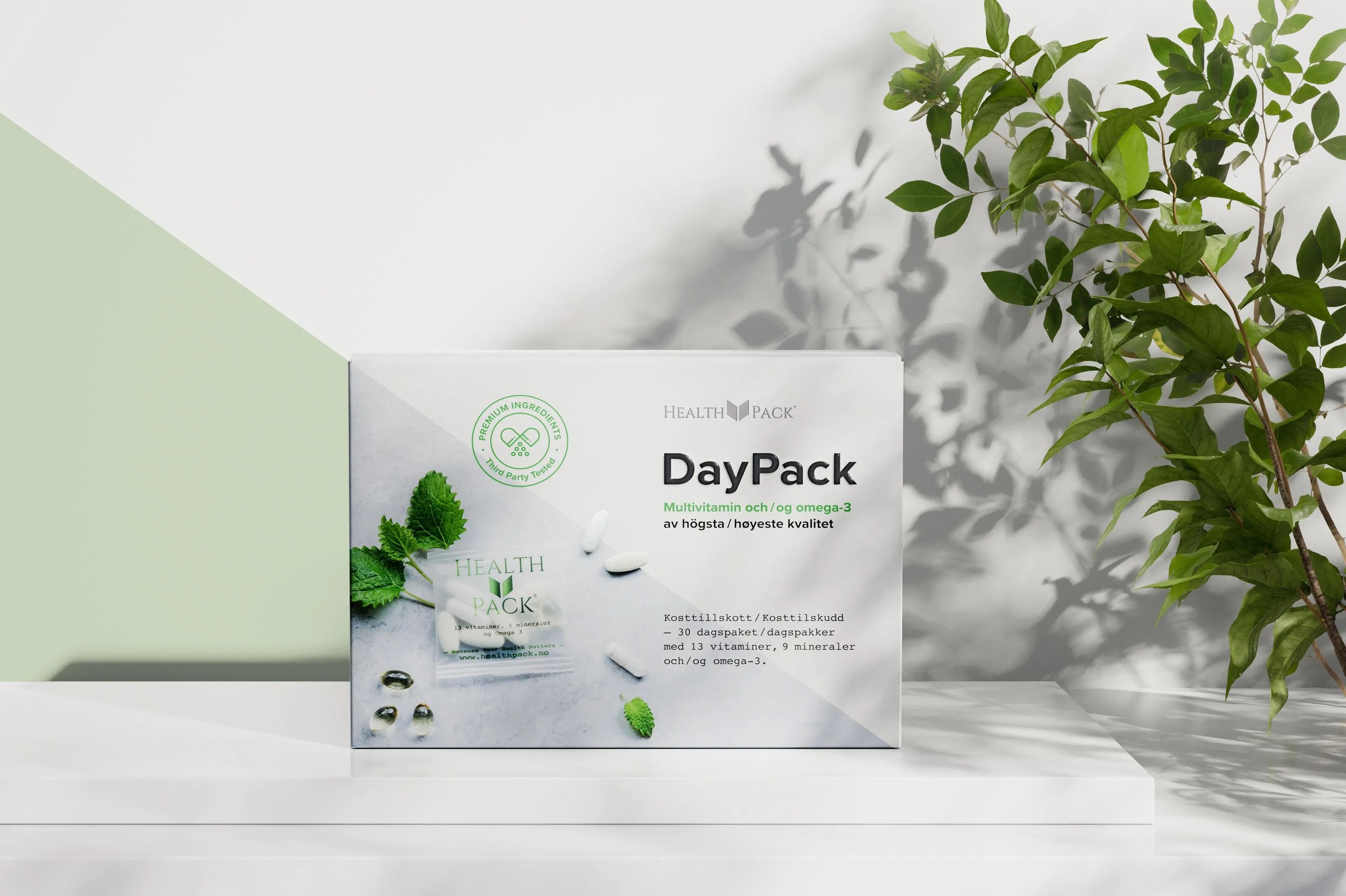 Box of Health Pack DayPack multivitamin and omega-3 supplements on a white shelf with green leaves and shadows in the background.