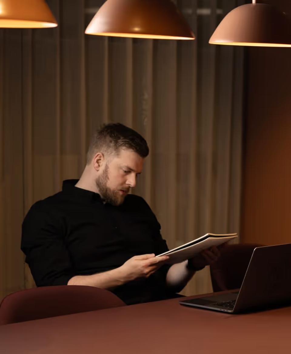 Jon looking concentrated. He has short hair and beard reading a document while sitting at a table with a laptop under warm pendant lights.