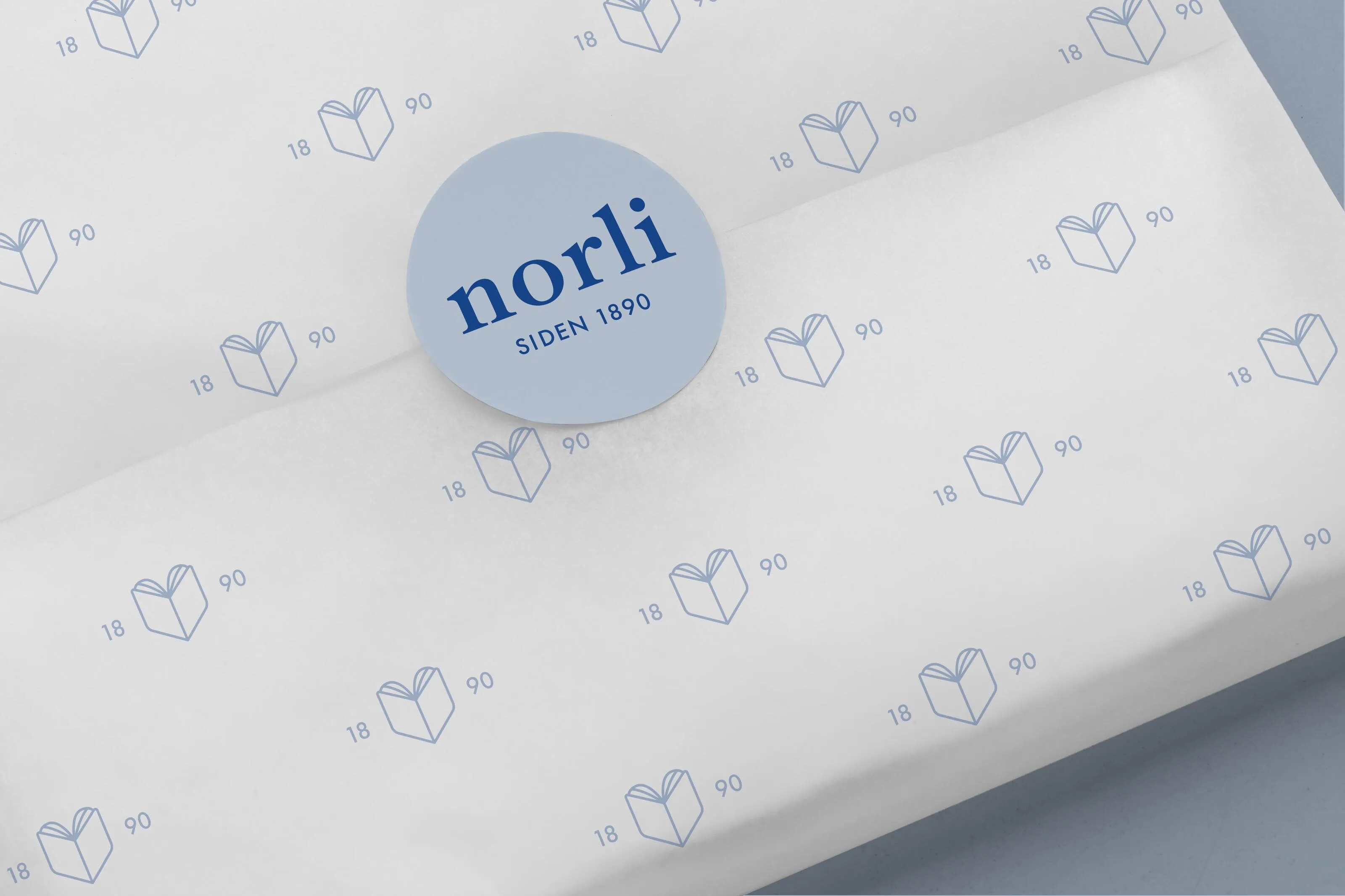 Wrapped package with white paper printed with blue book icons and numbers 18 and 90, sealed with a blue circular sticker labelled 'norli SIDEN 1890'.