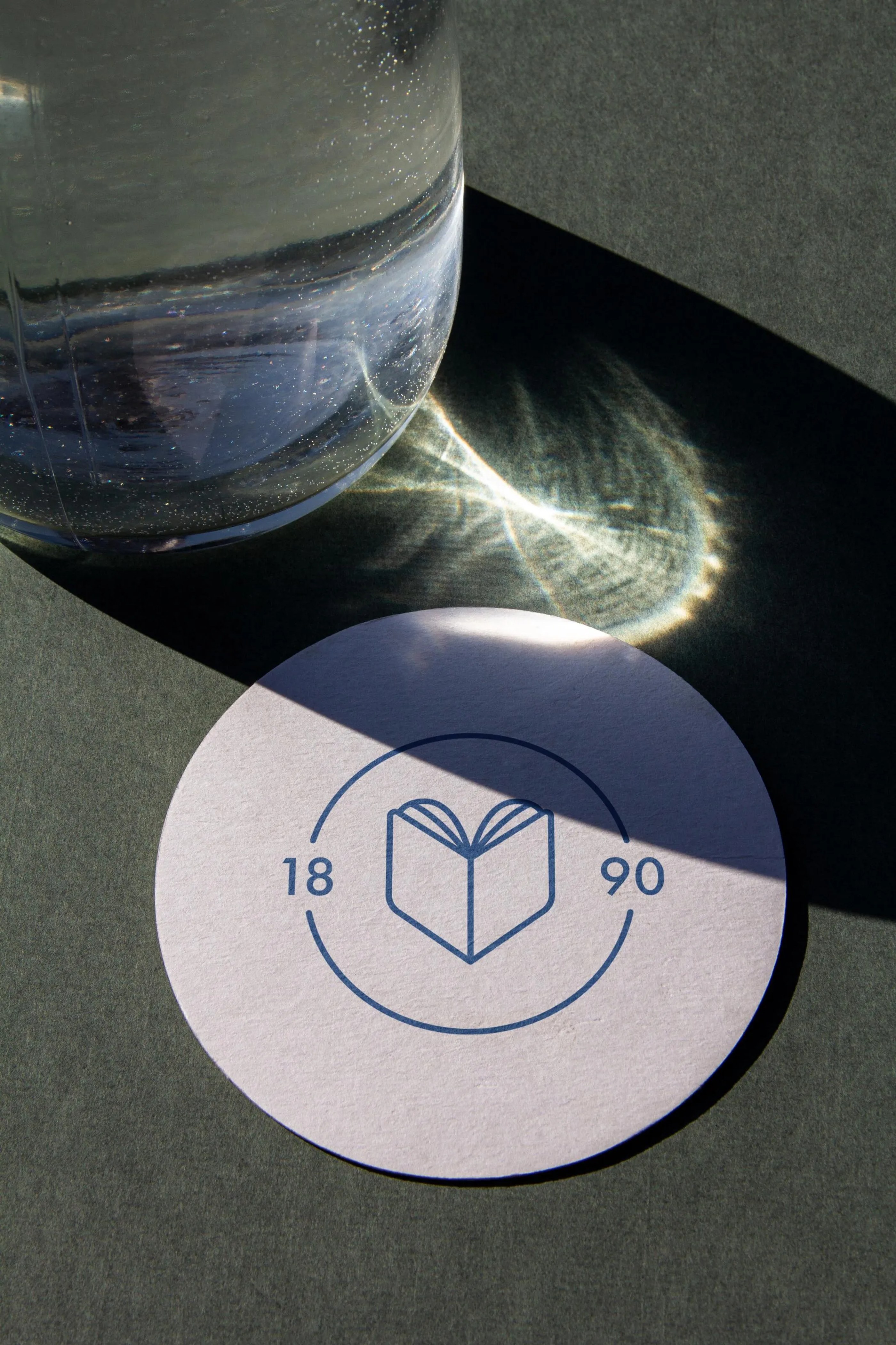 Close-up of a round white coaster with an open book logo and the numbers 18 and 90 on a dark surface, partially shaded by a glass of water casting light reflections.