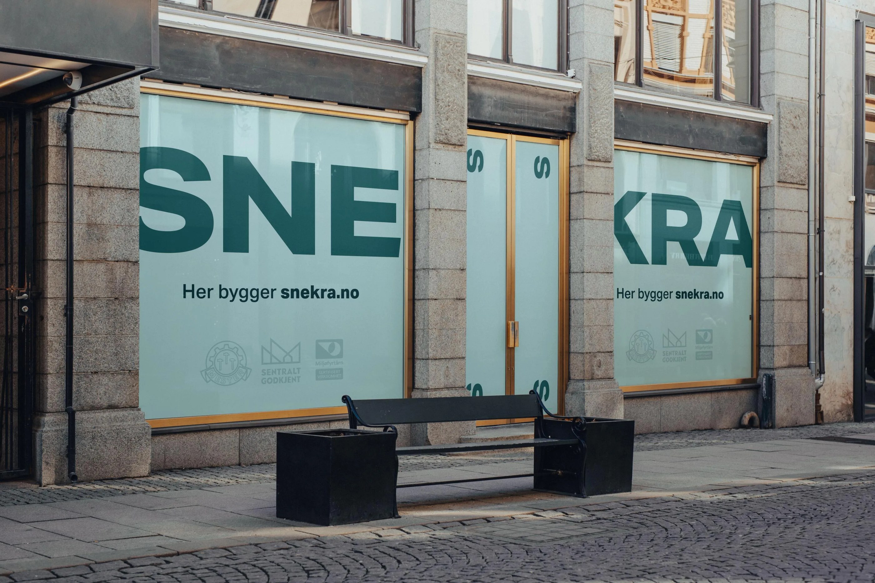 Storefront with large windows and gold-trimmed doors covered with signage reading 'SNE' and 'KRA' and text 'Her bygger snekra.no', with a black bench in front on cobblestone pavement.