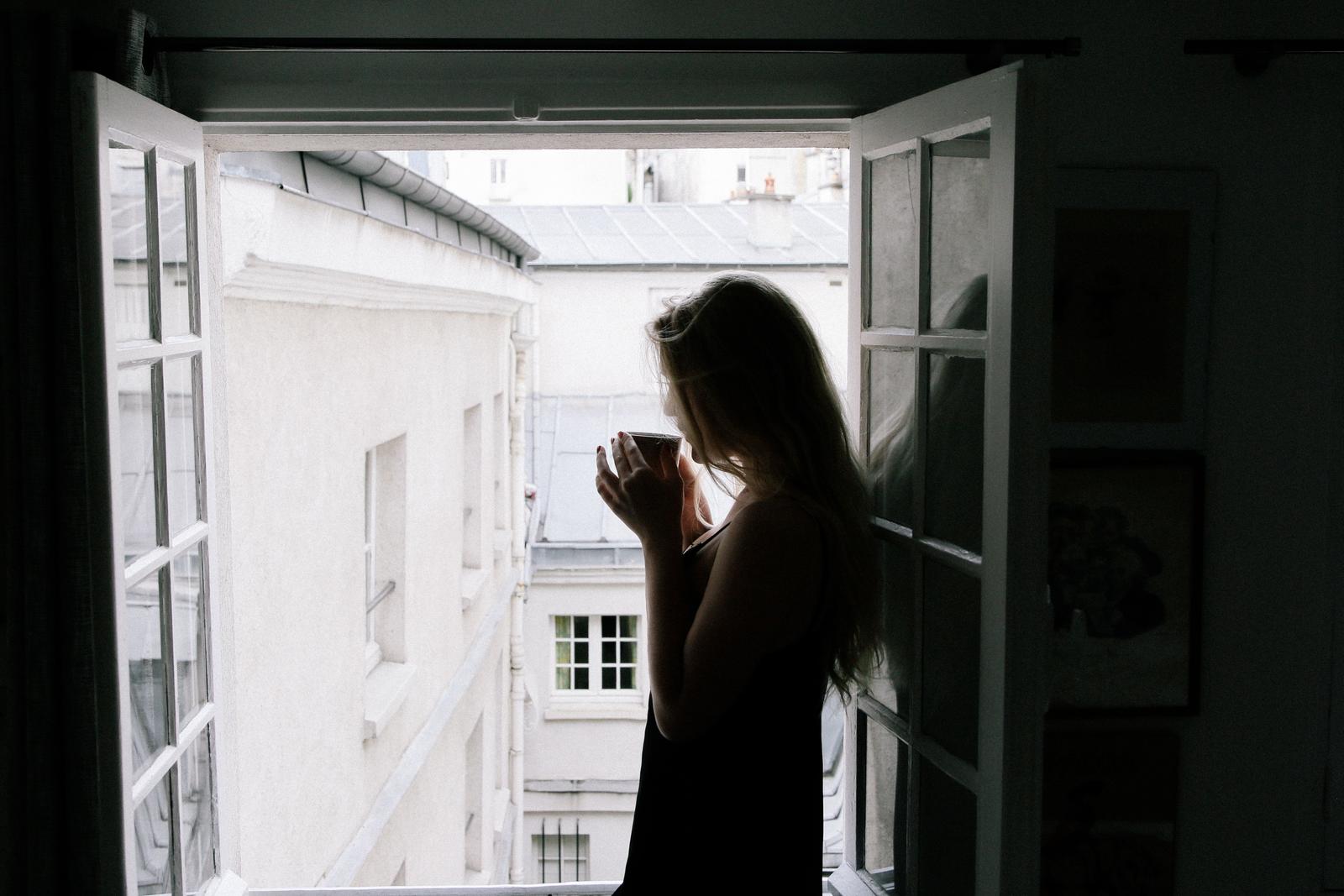Silhouet van een vrouw met een kop koffie voor een raam, ochtendlicht over de stad